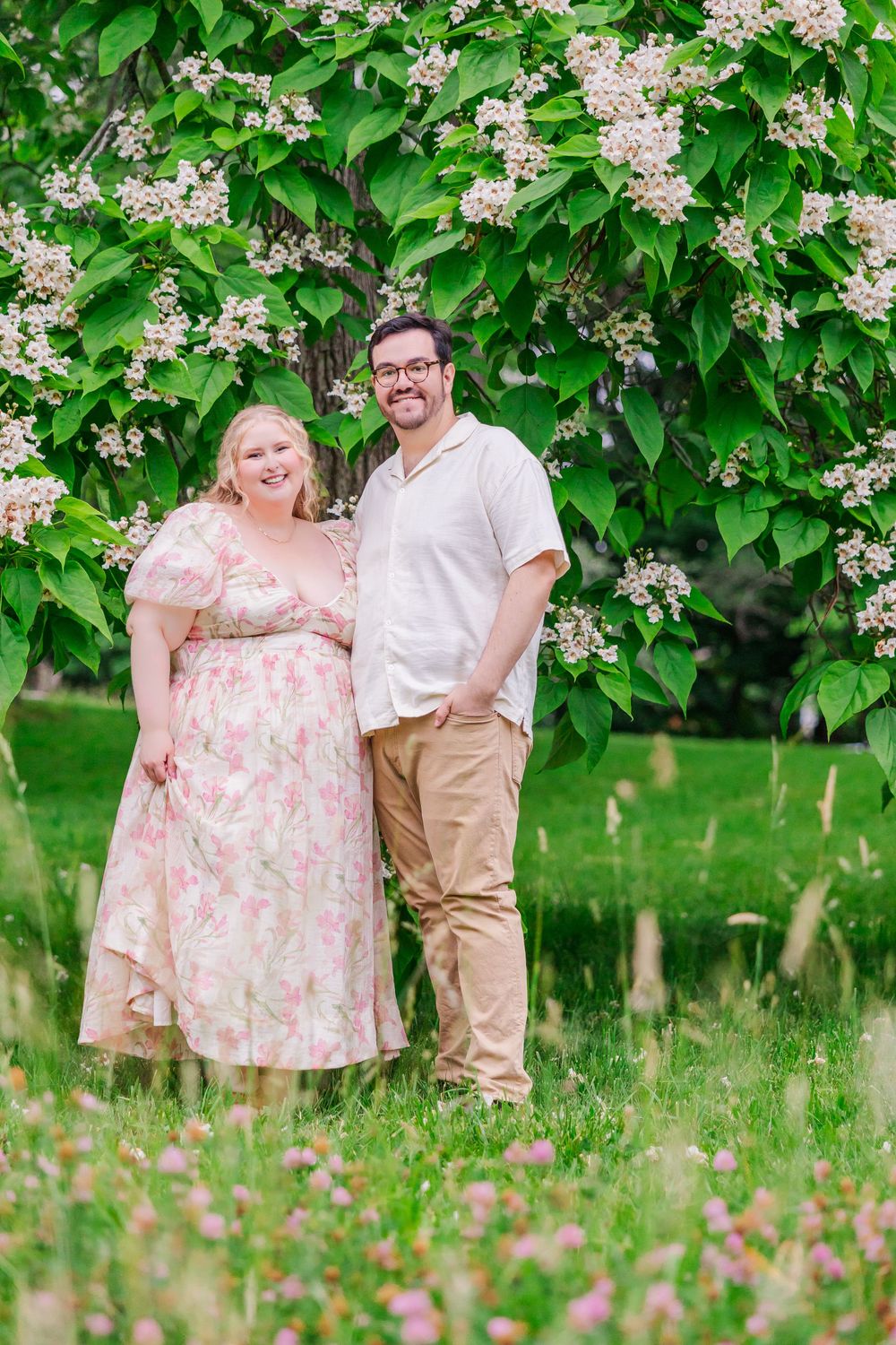 A couple poses together beneath blooming white lilac bushes in a spring garden setting.