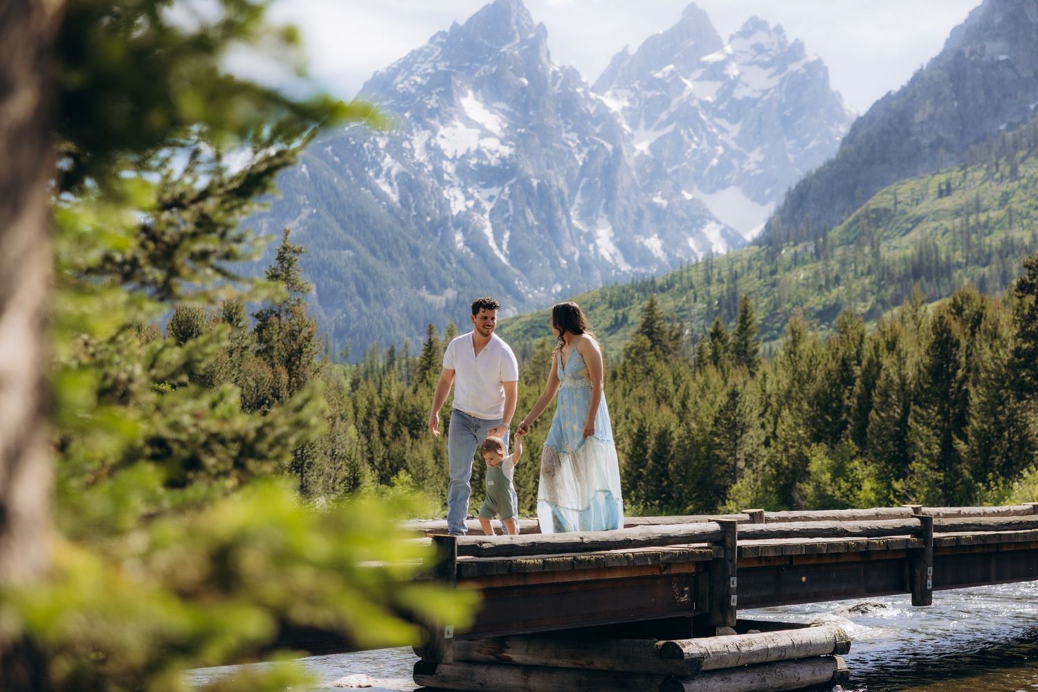 A couple stands on a wooden dock overlooking a scenic mountain vista with snow-capped peaks and evergreen forest.