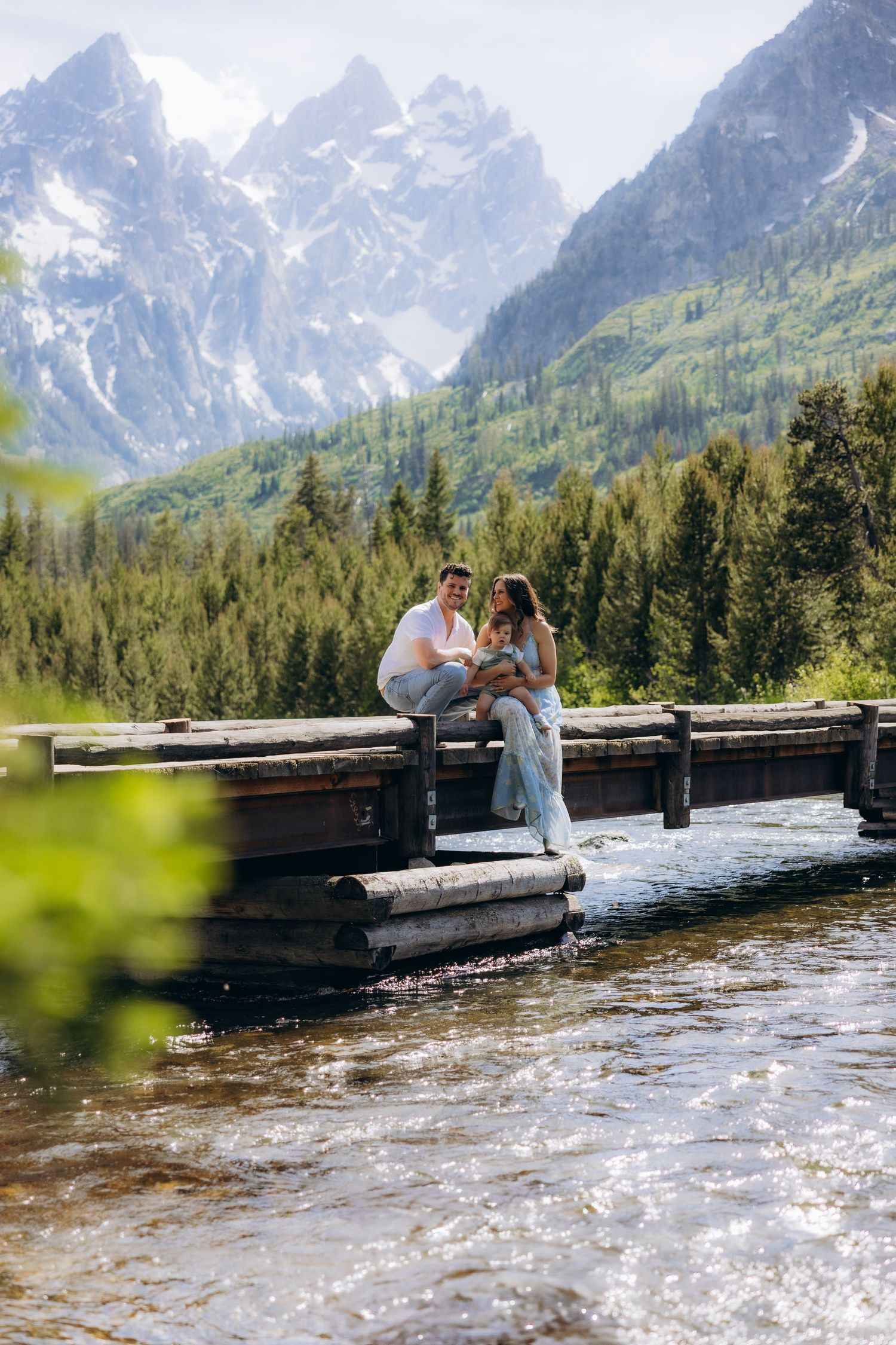 A couple shares an intimate moment on a wooden boardwalk against a stunning mountain backdrop with pine forests.