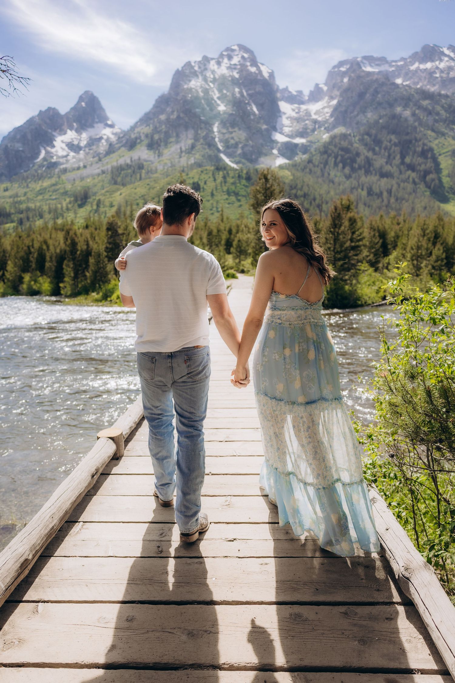 Couple holding hands while walking on wooden bridge with scenic mountain backdrop and river in Grand Teton National Park.
