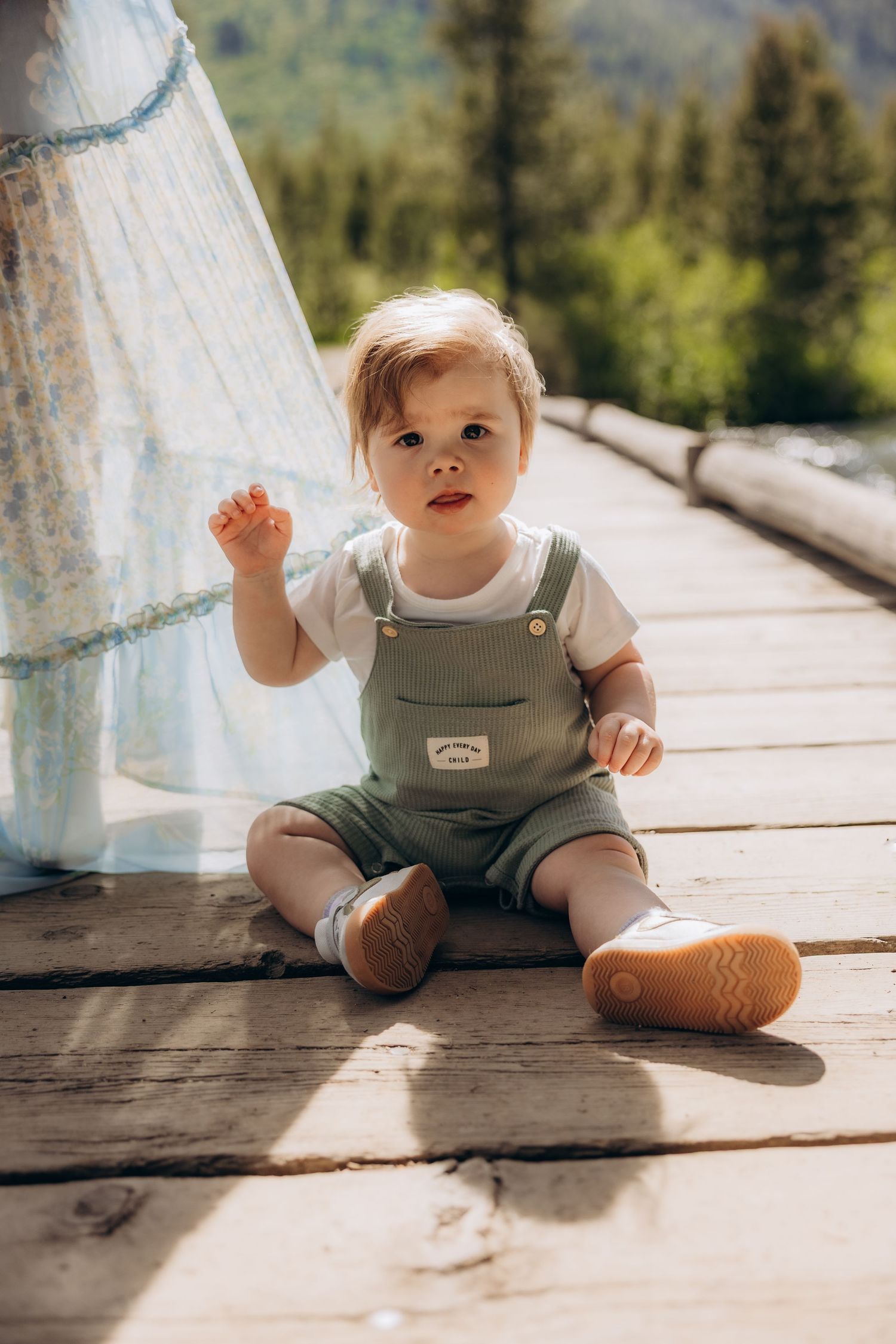 Small child sits on wooden deck wearing green overalls and brown shoes in outdoor mountain setting.