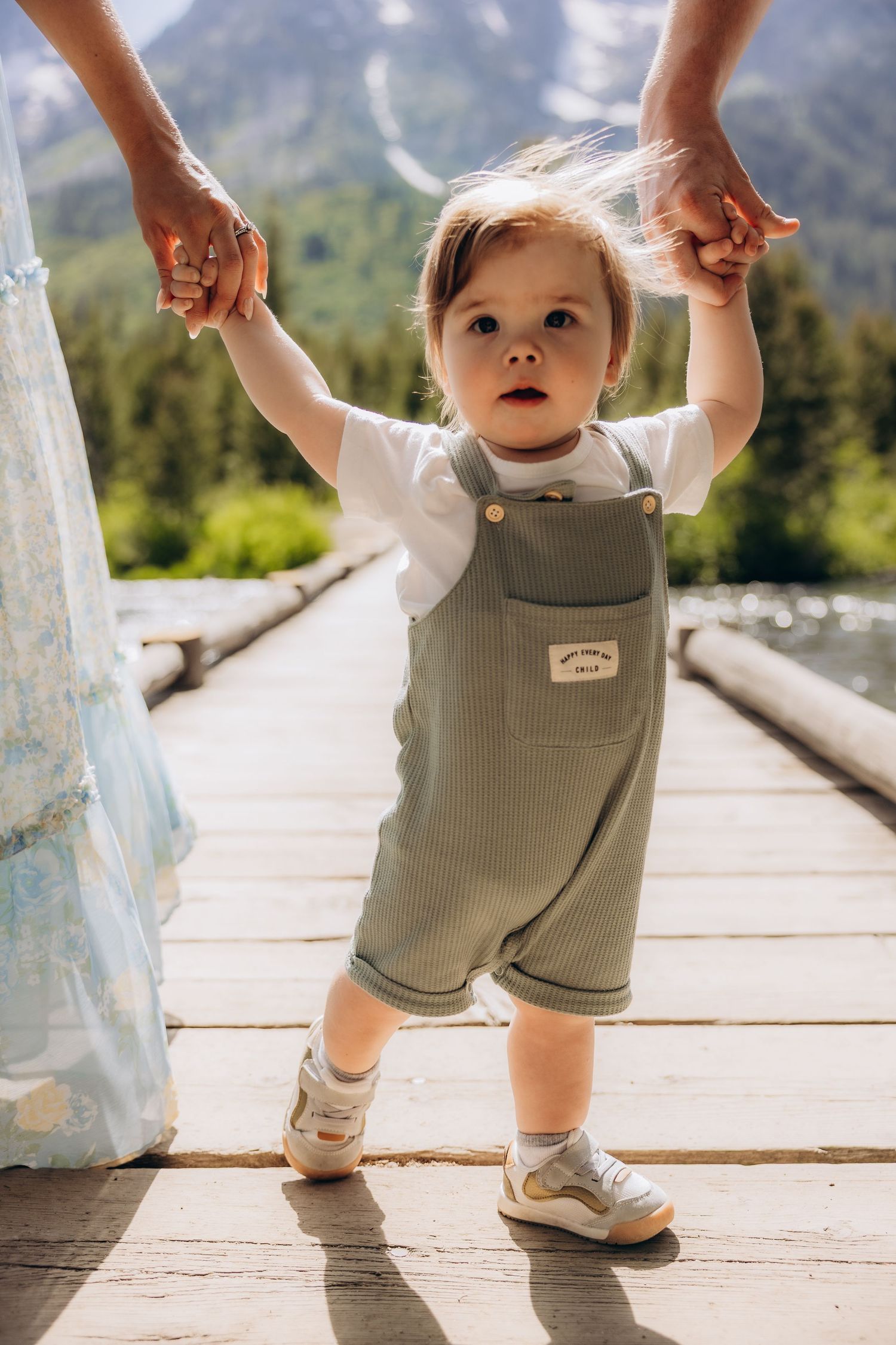 Young toddler in overalls taking first steps on wooden bridge with mountains in background.