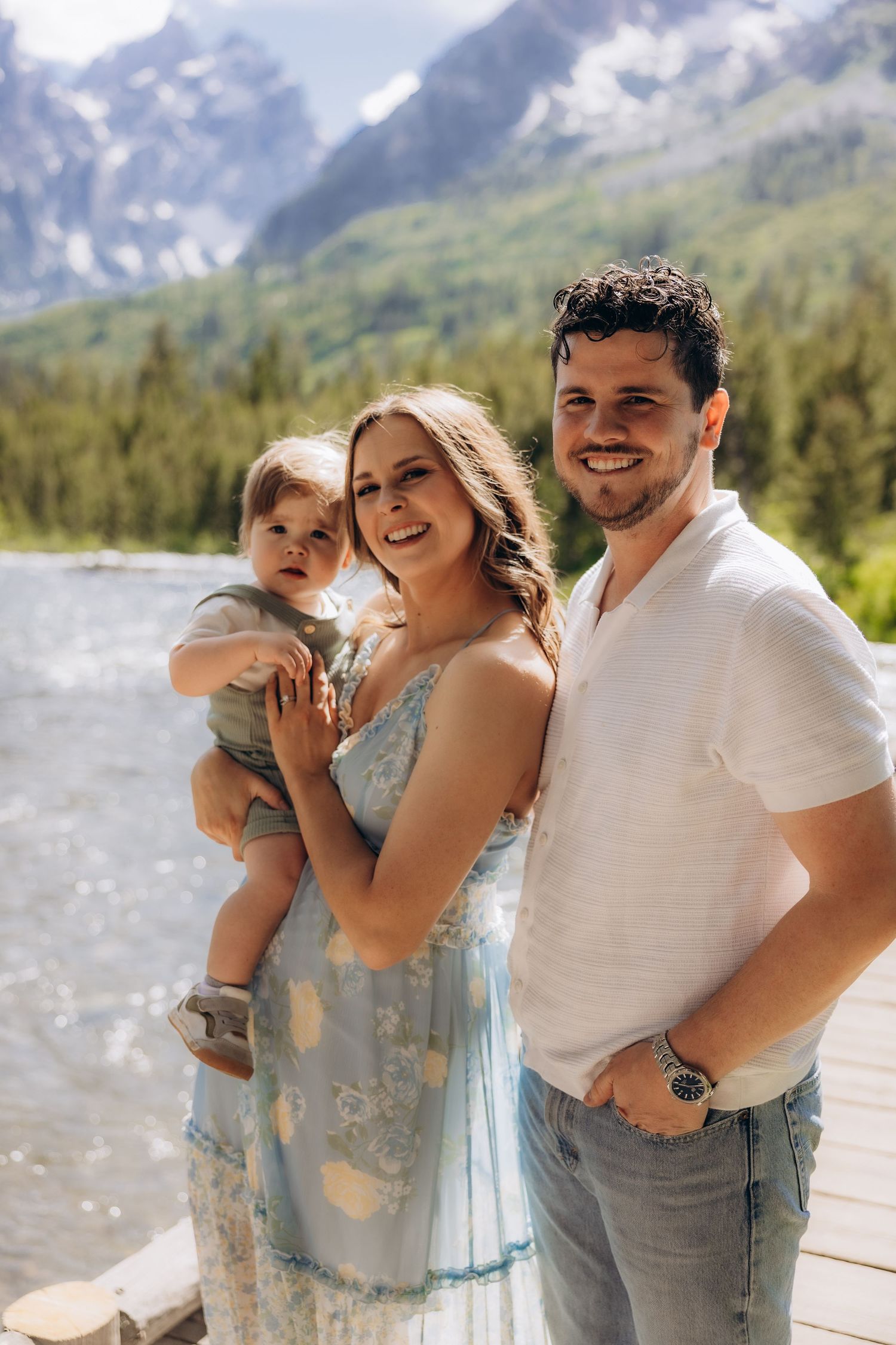 Family of three posing together on wooden bridge with snow-capped mountains and forest in background.