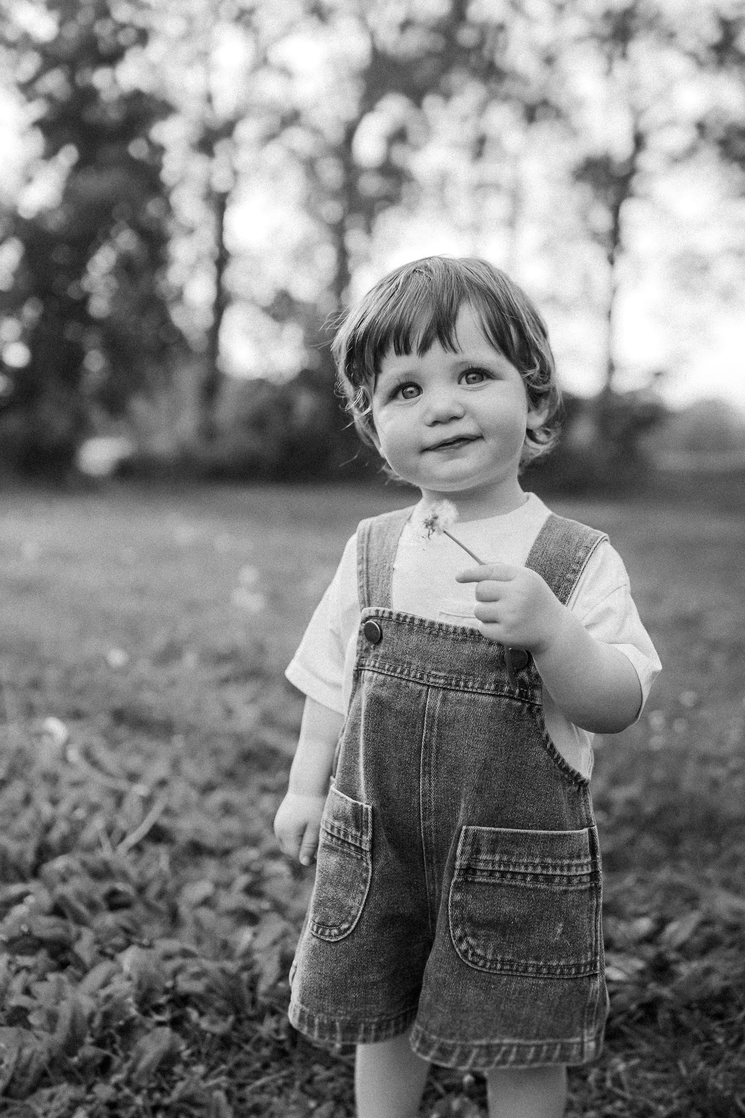 A small child wearing denim overalls stands in a grassy outdoor setting in black and white photography.