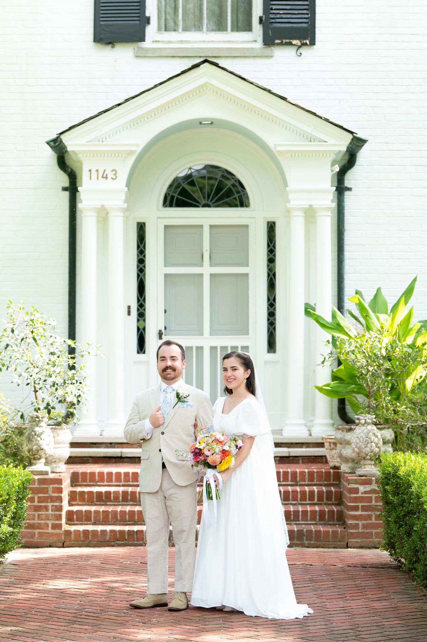 Wedding couple poses in front of a classic white Southern home with green shutters and brick steps.