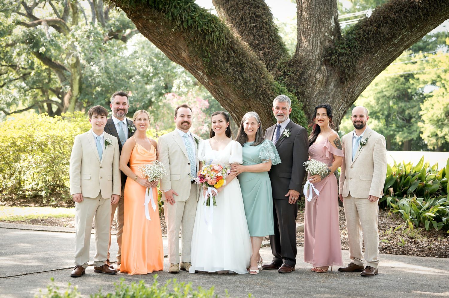 Large wedding party in pastel dresses and light suits poses beneath sprawling oak trees.