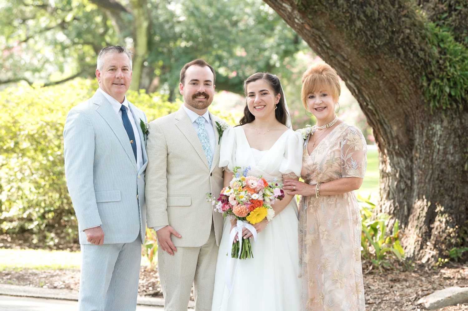 Wedding party members gather under oak trees wearing pastel colors and light suits.