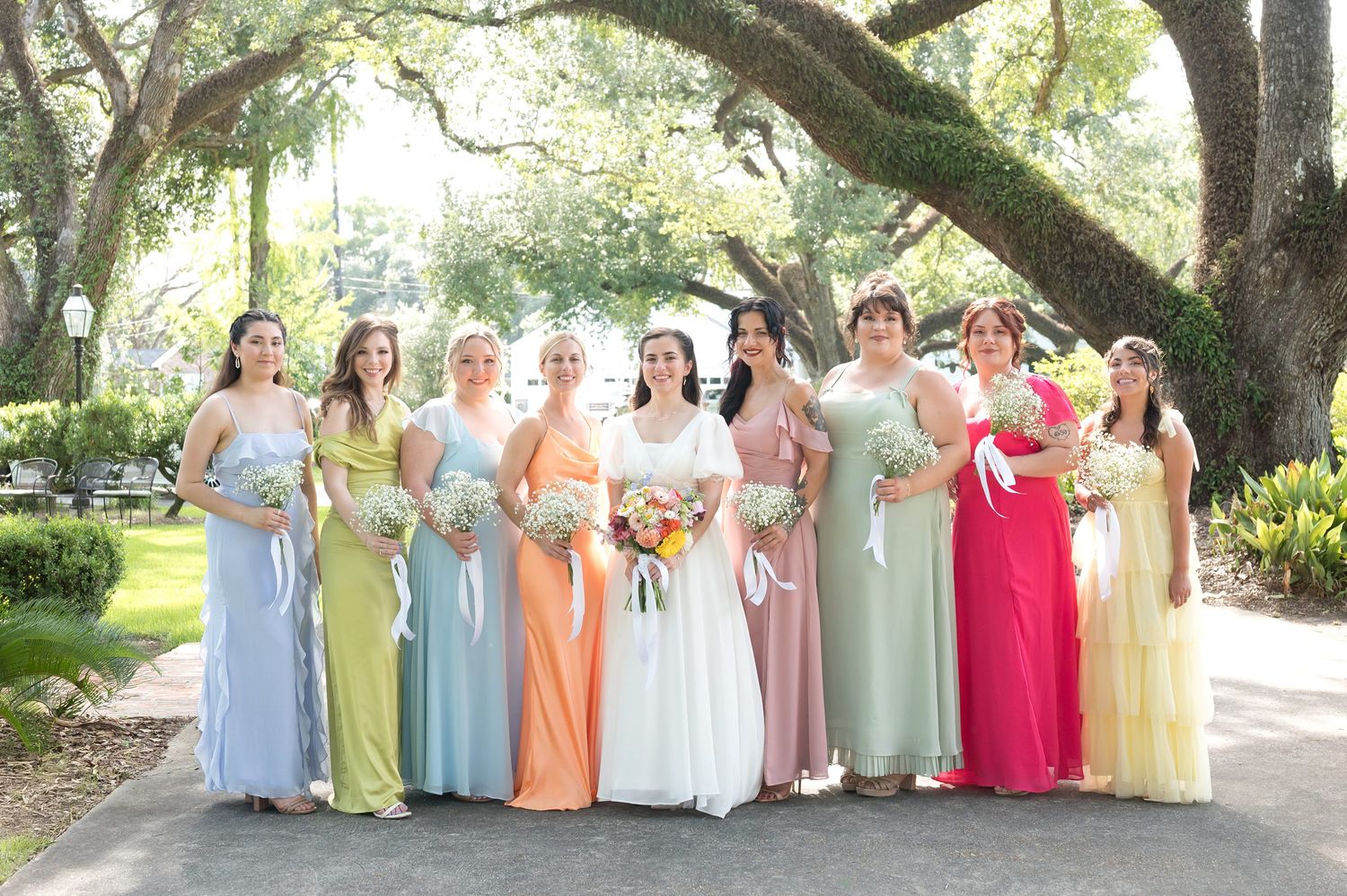 Wedding party poses under large oak trees wearing pastel colored dresses and light suits.
