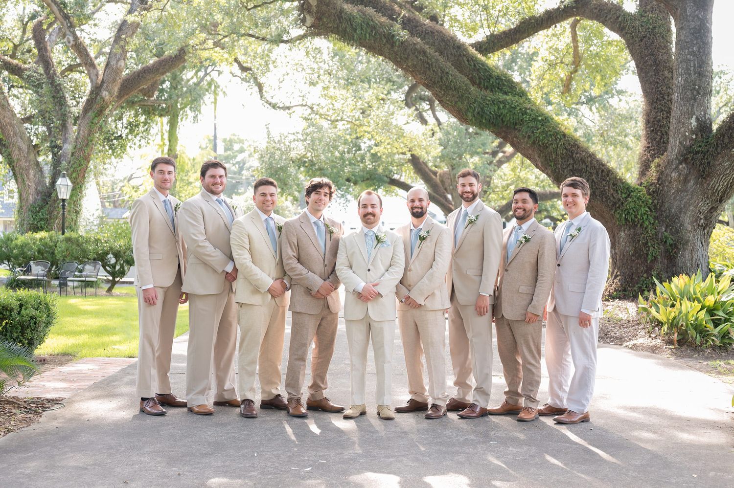 Groomsmen in light colored suits pose together under shady trees.