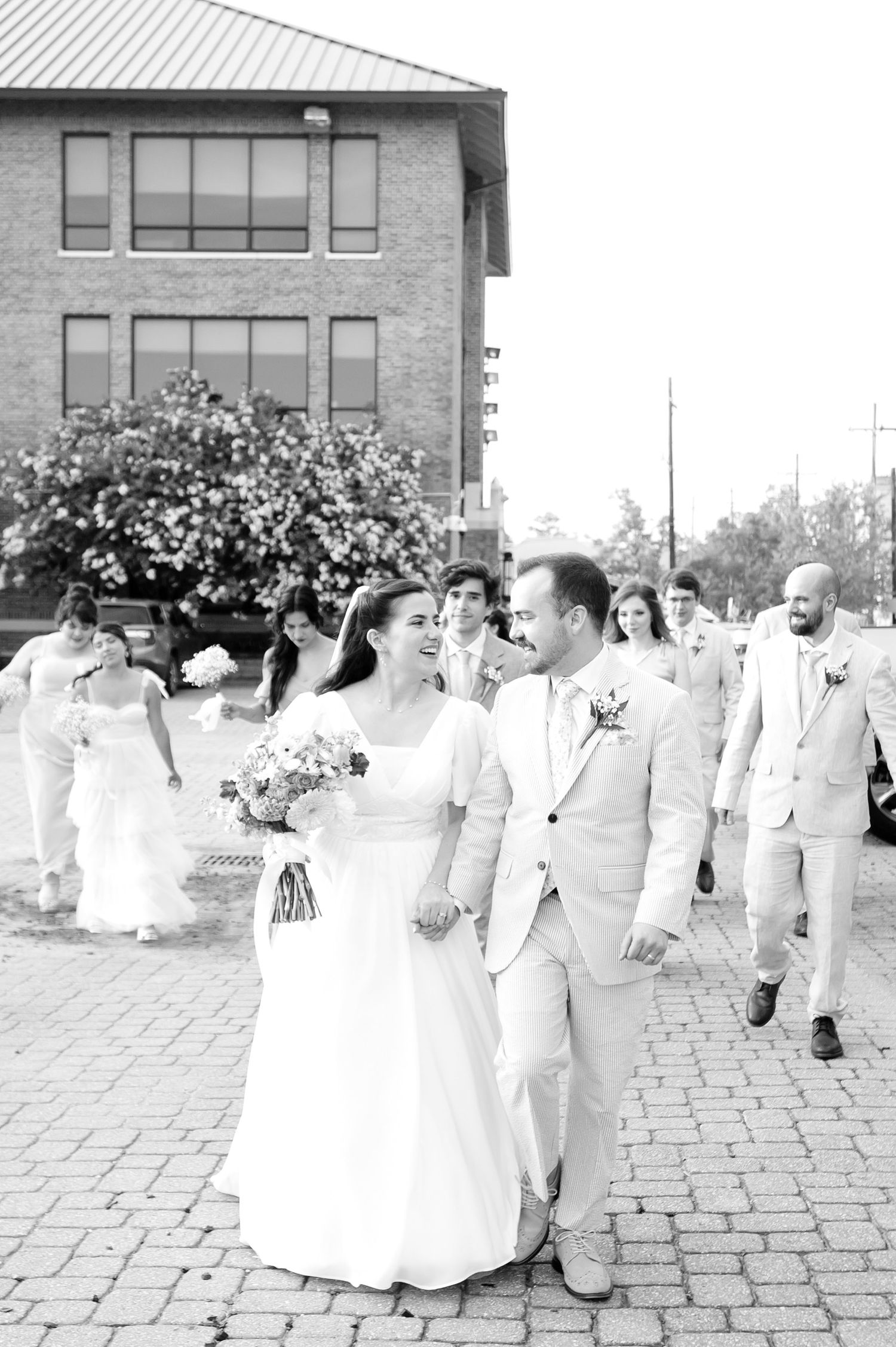 Wedding party walking along a brick pathway outside a modern building in black and white.