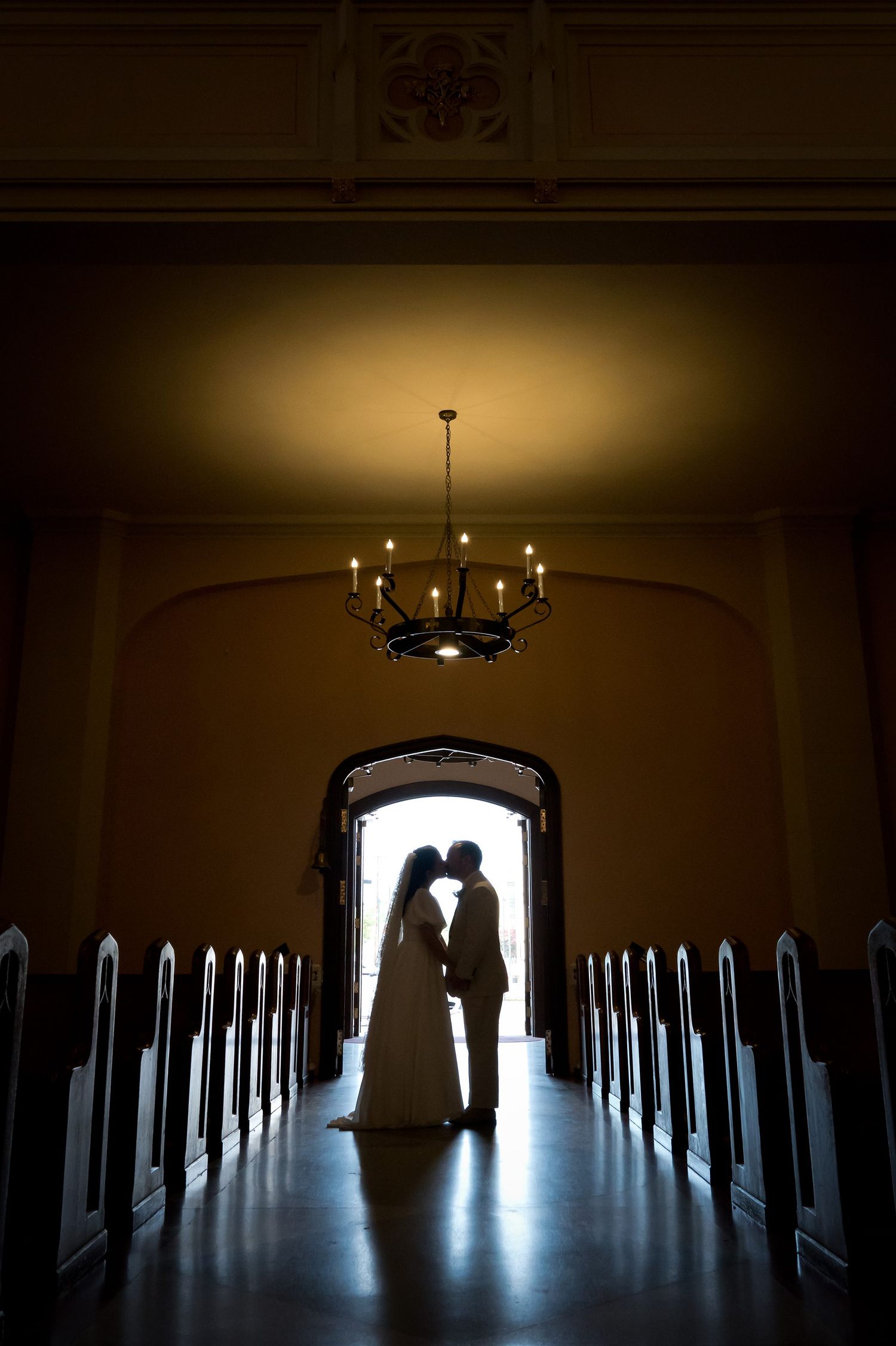 Silhouette of couple in church doorway with dramatic backlighting and chandelier above.