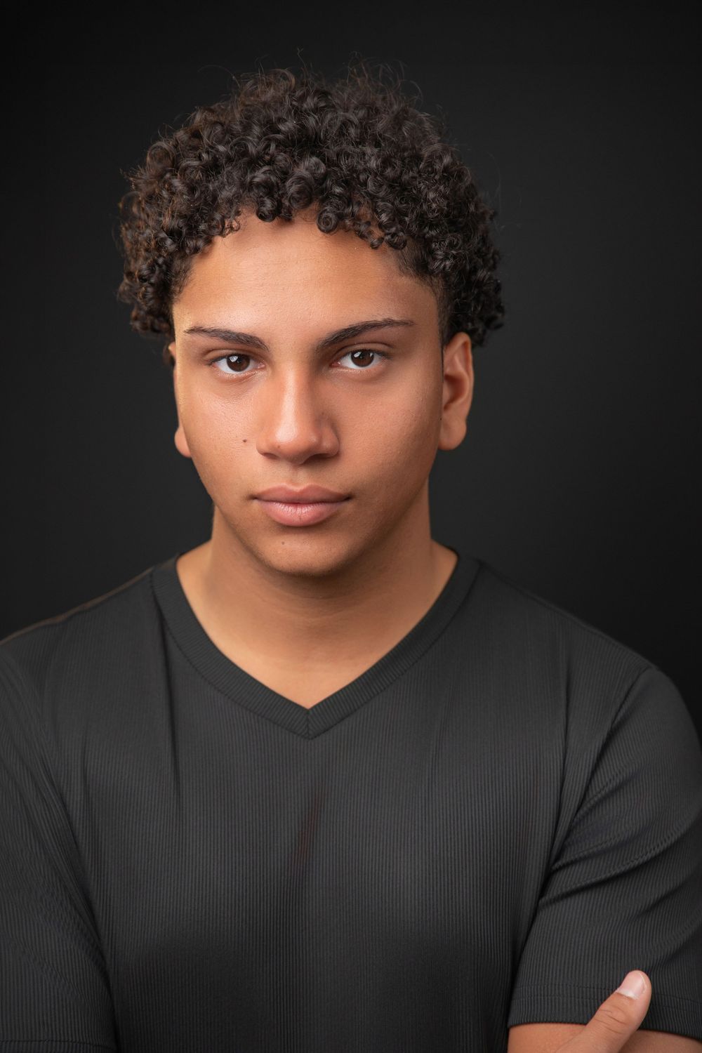 Young man with curly hair wearing a black v-neck shirt, arms crossed, posing against a dark background.