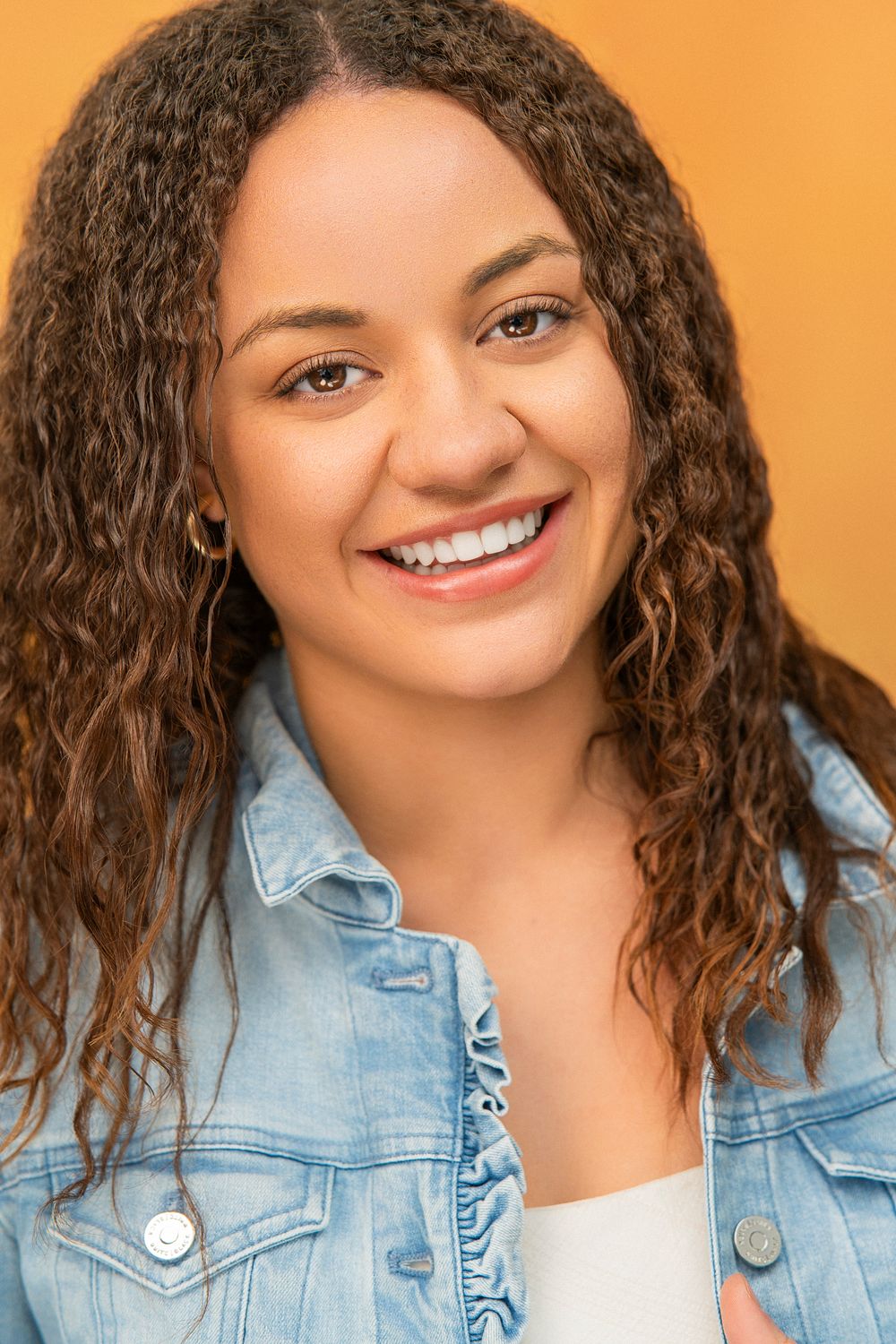 Smiling person with curly brown hair wearing a light denim jacket against an orange background.