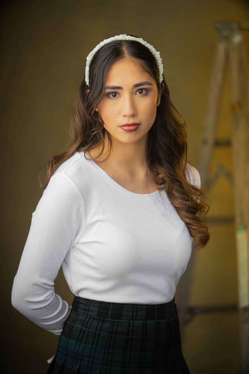 Portrait of someone in a white long-sleeved top and plaid skirt with wavy brown hair and a delicate headband.