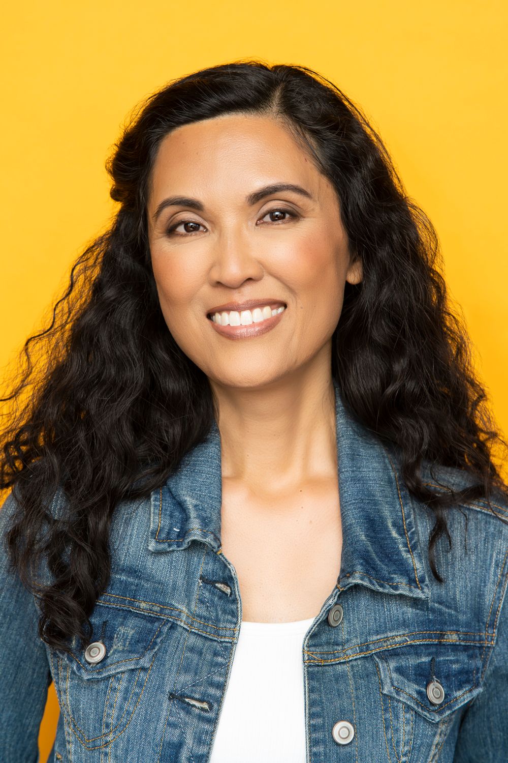Professional portrait of someone with long dark curly hair wearing a denim jacket against a yellow background.
