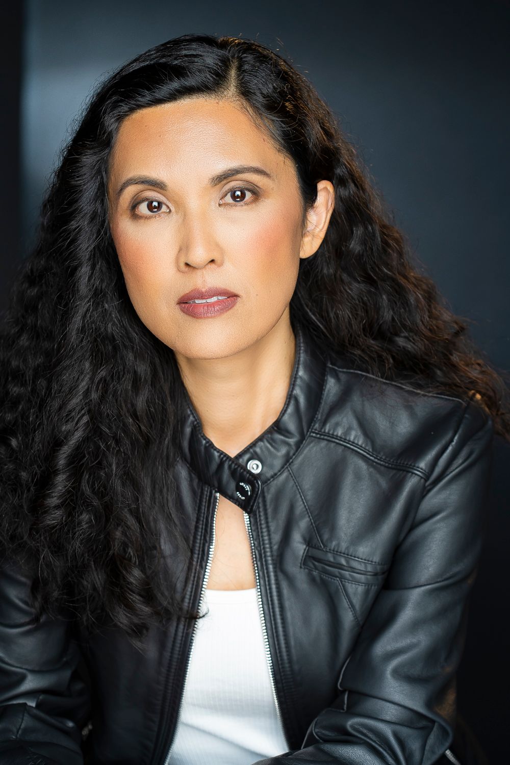 Professional portrait of someone with long dark wavy hair wearing a black leather jacket and white top against dark background.