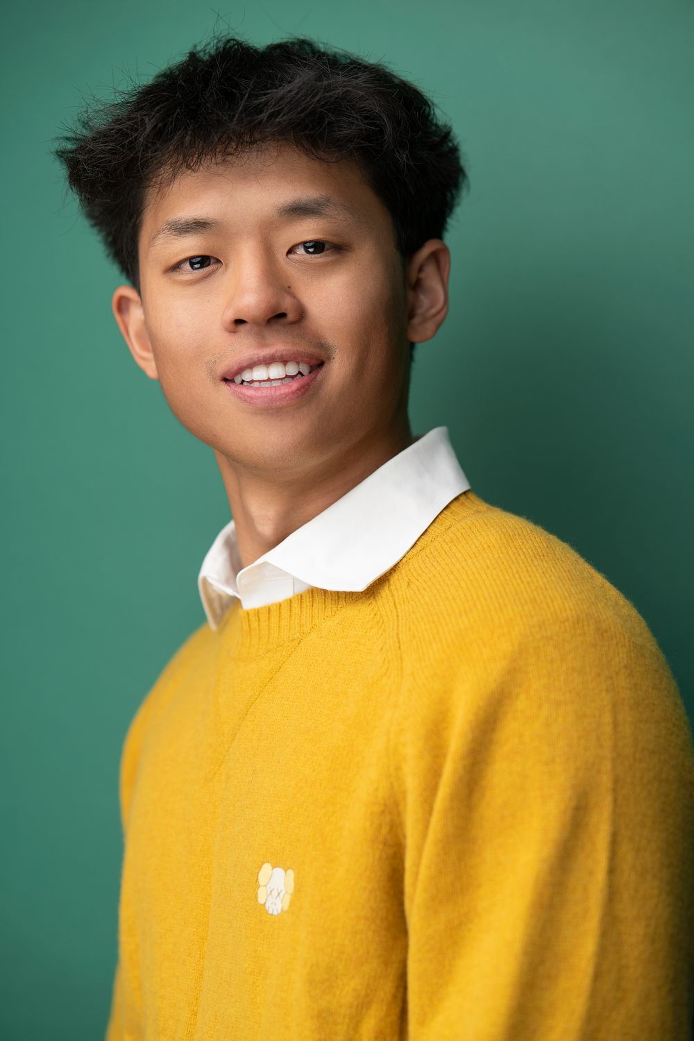Portrait of someone in a bright yellow sweater and white collar against a teal background.