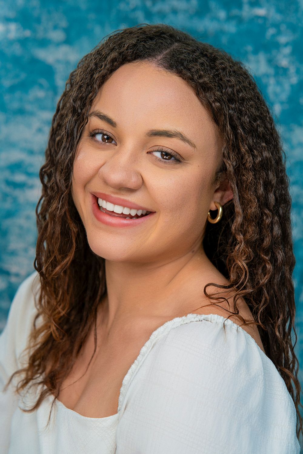 A person with curly brown hair and a bright smile wearing a white top against a blue background.