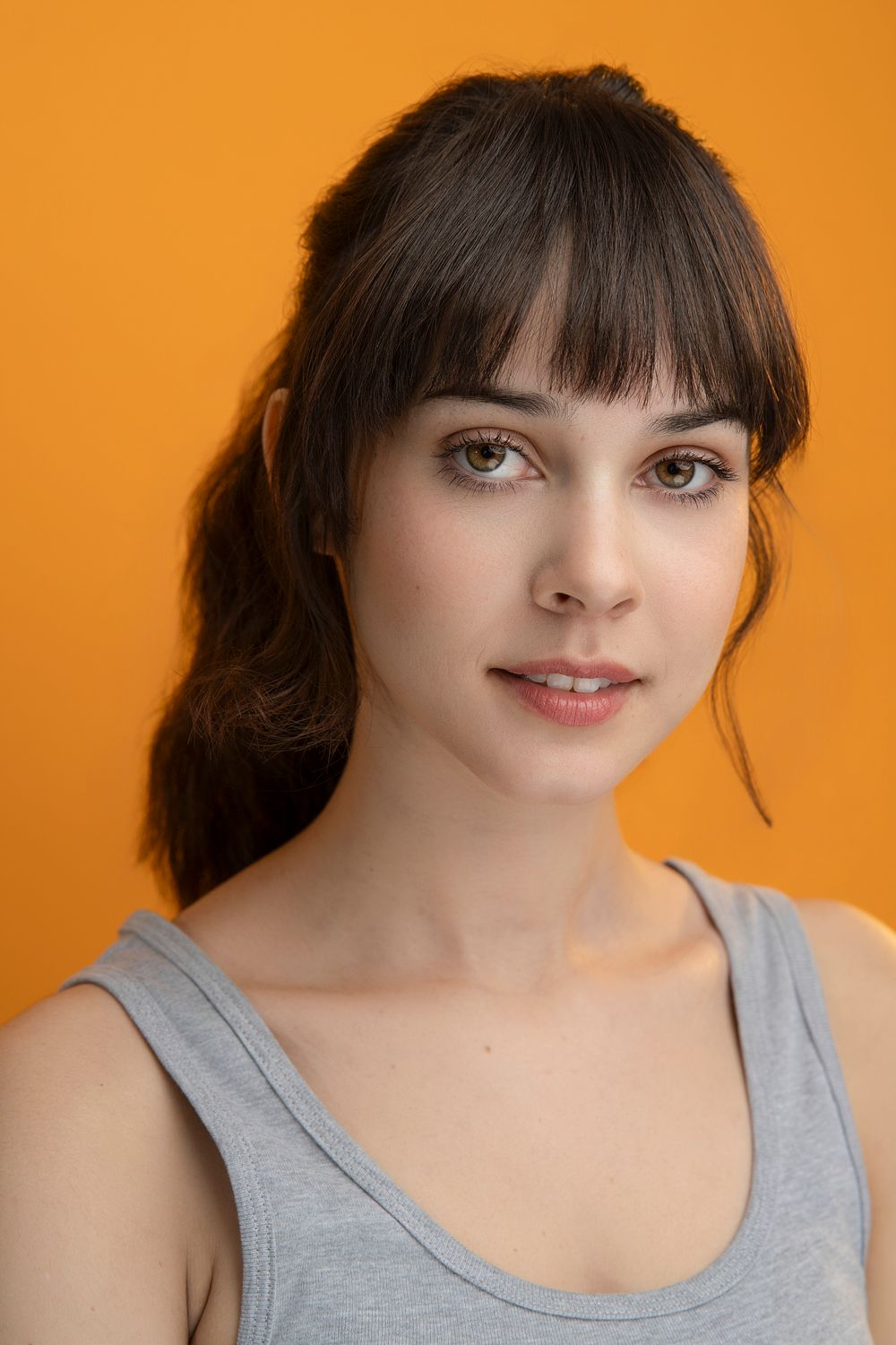 Portrait against orange background showing warm smile and bangs framing the face in casual grey tank top.