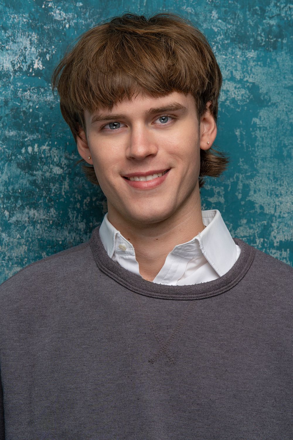 Portrait against a textured turquoise wall wearing a gray sweater over white collared shirt.