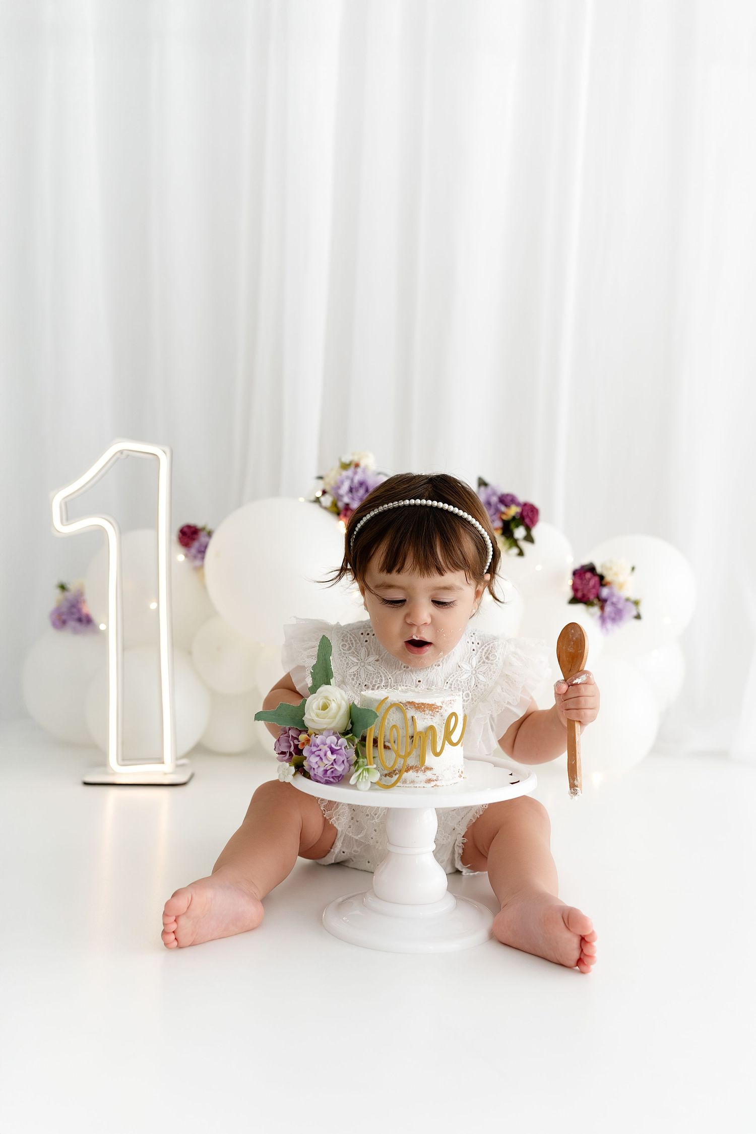 A first birthday celebration setup with a white cake on a stand, large number one light, and purple floral decorations.
