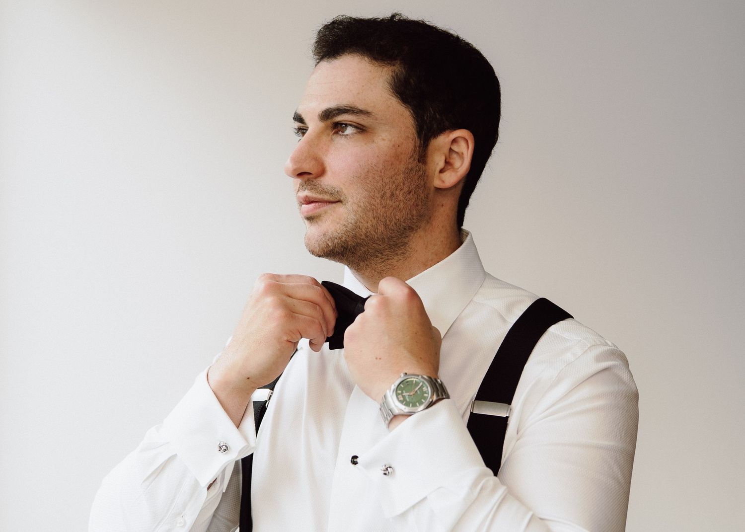 Groom in white dress shirt and suspenders adjusts his bow tie while getting ready.
