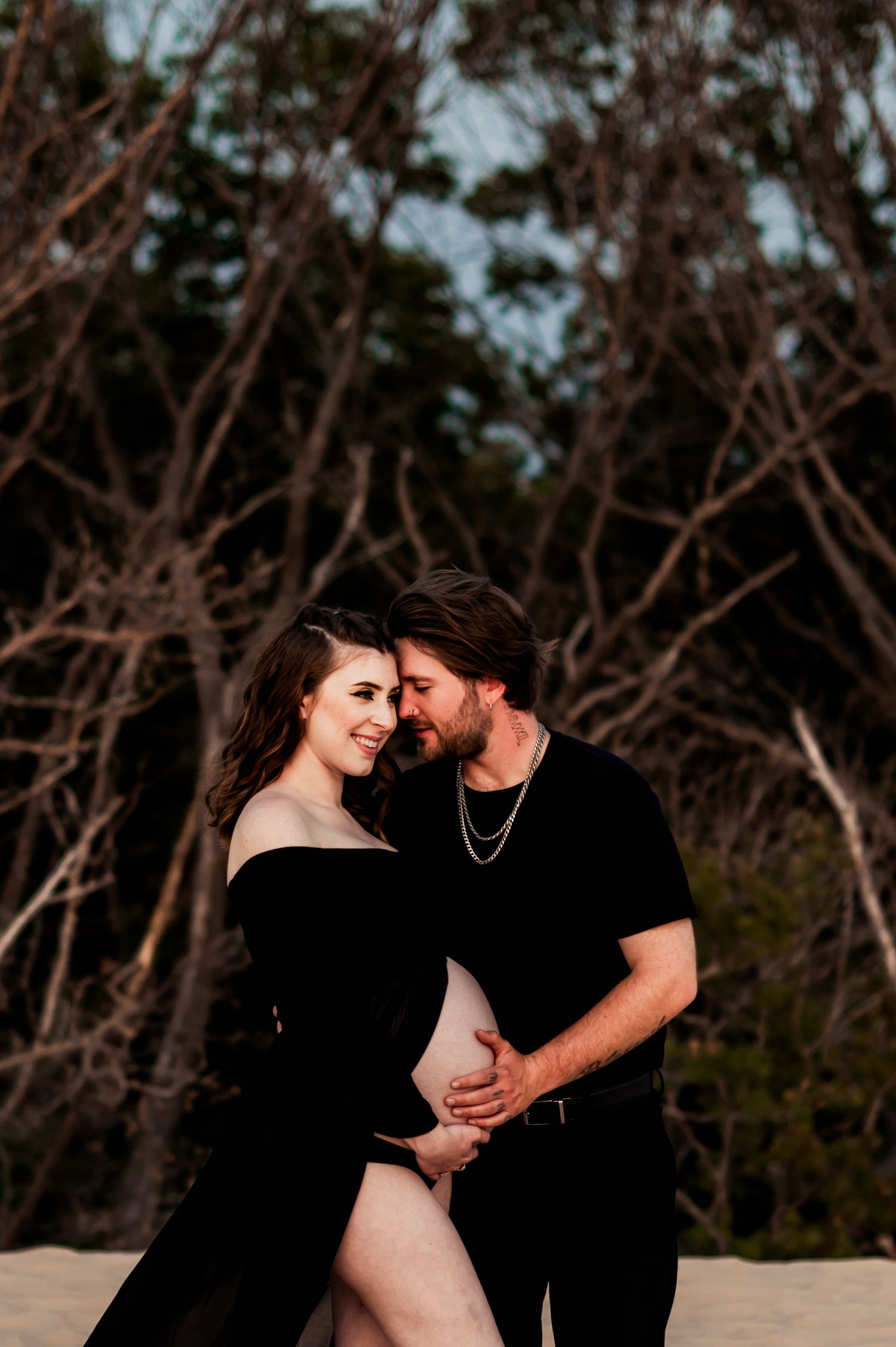 Romantic maternity couple in black clothing embrace against rustic wooded backdrop during sunset photo session.