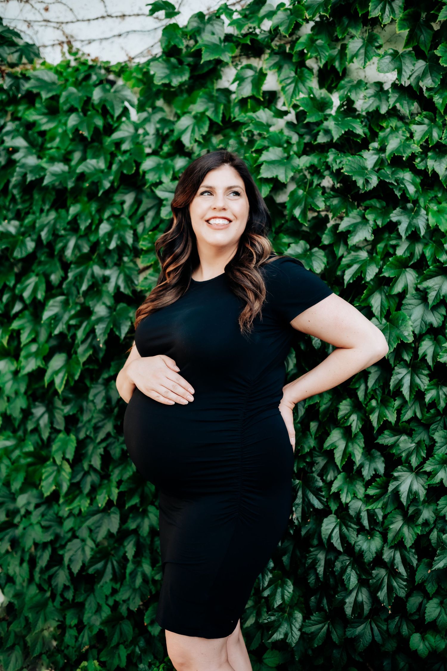 A pregnant woman in a black dress smiles while standing against a lush green ivy-covered wall.
