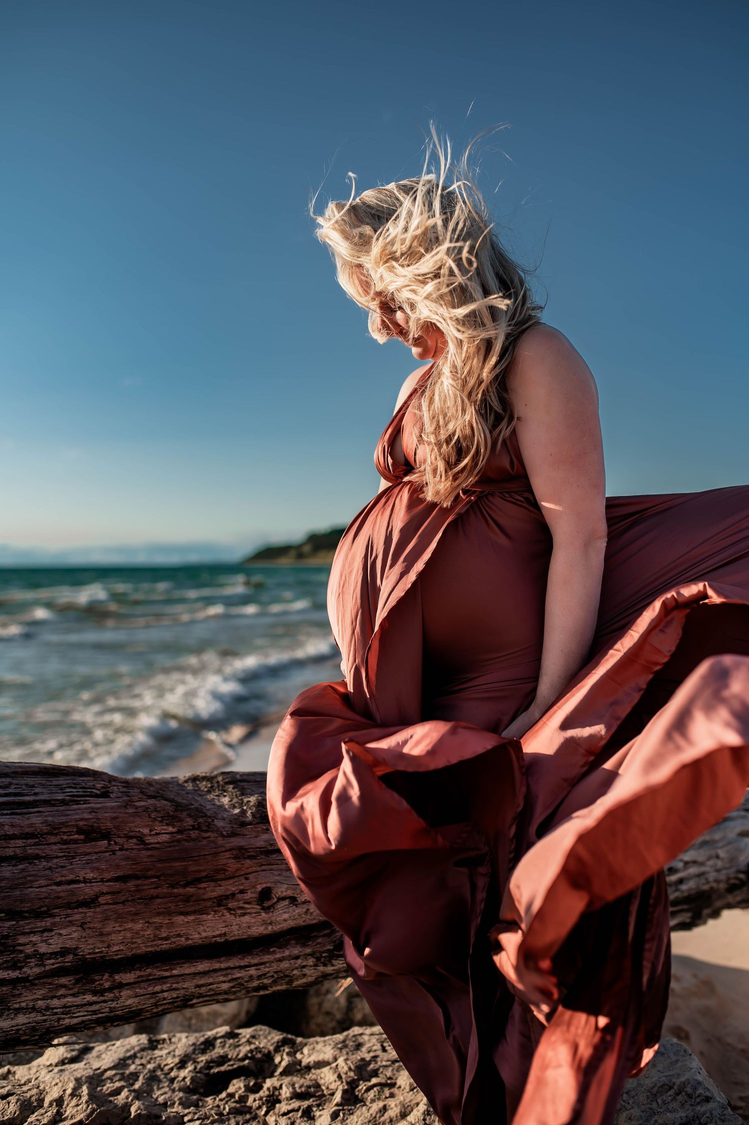 A flowing rust-colored dress billows in the ocean breeze as someone stands on rocky coastal outcrop at sunset.