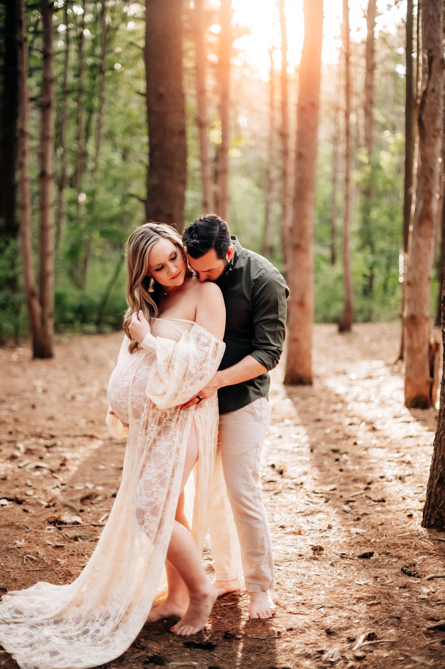 A romantic forest maternity photo featuring an intimate moment between a couple surrounded by sunlit pine trees.