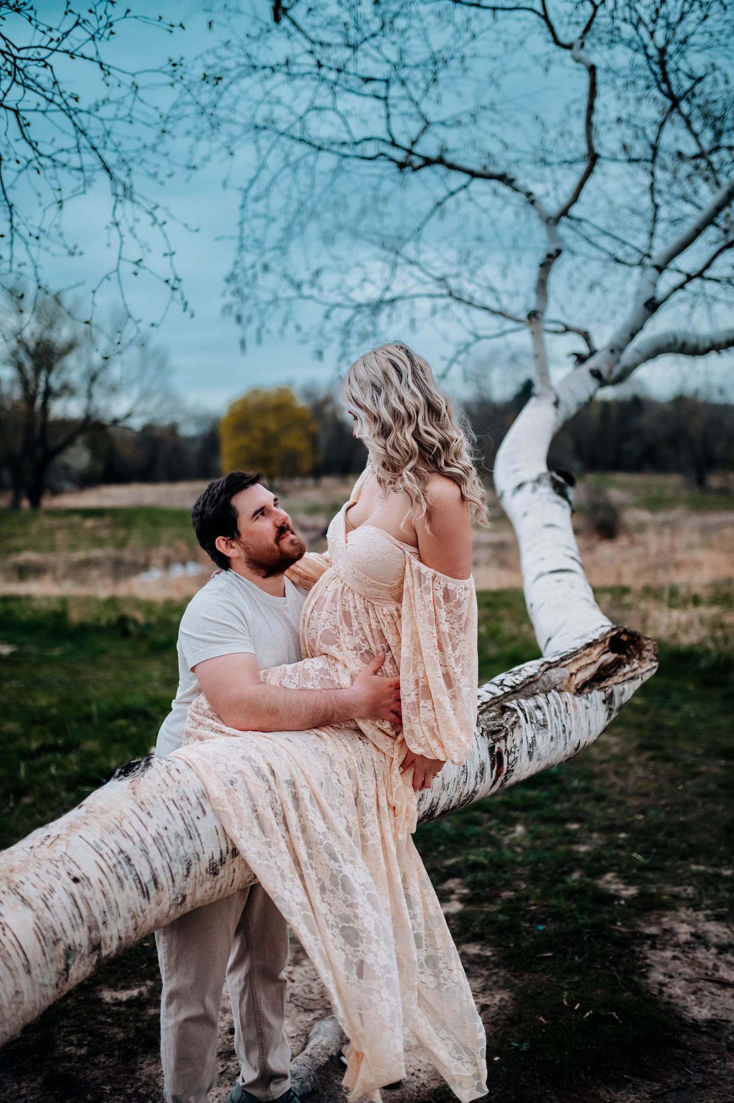 A romantic couple sits together on a fallen birch tree branch during a dreamy outdoor photoshoot in nature.
