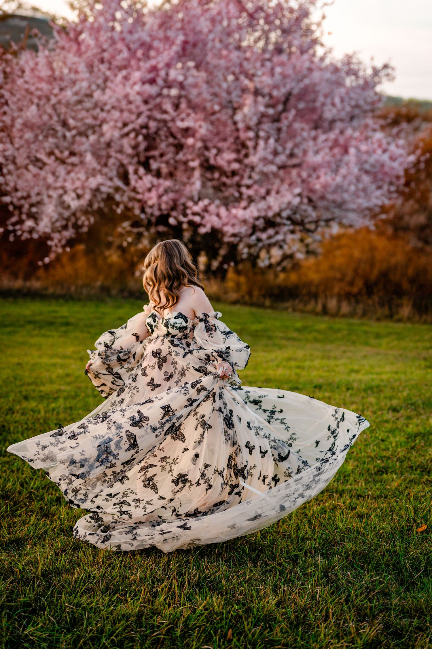 A person in a flowing white floral dress spins joyfully beneath blooming cherry blossom trees during springtime.