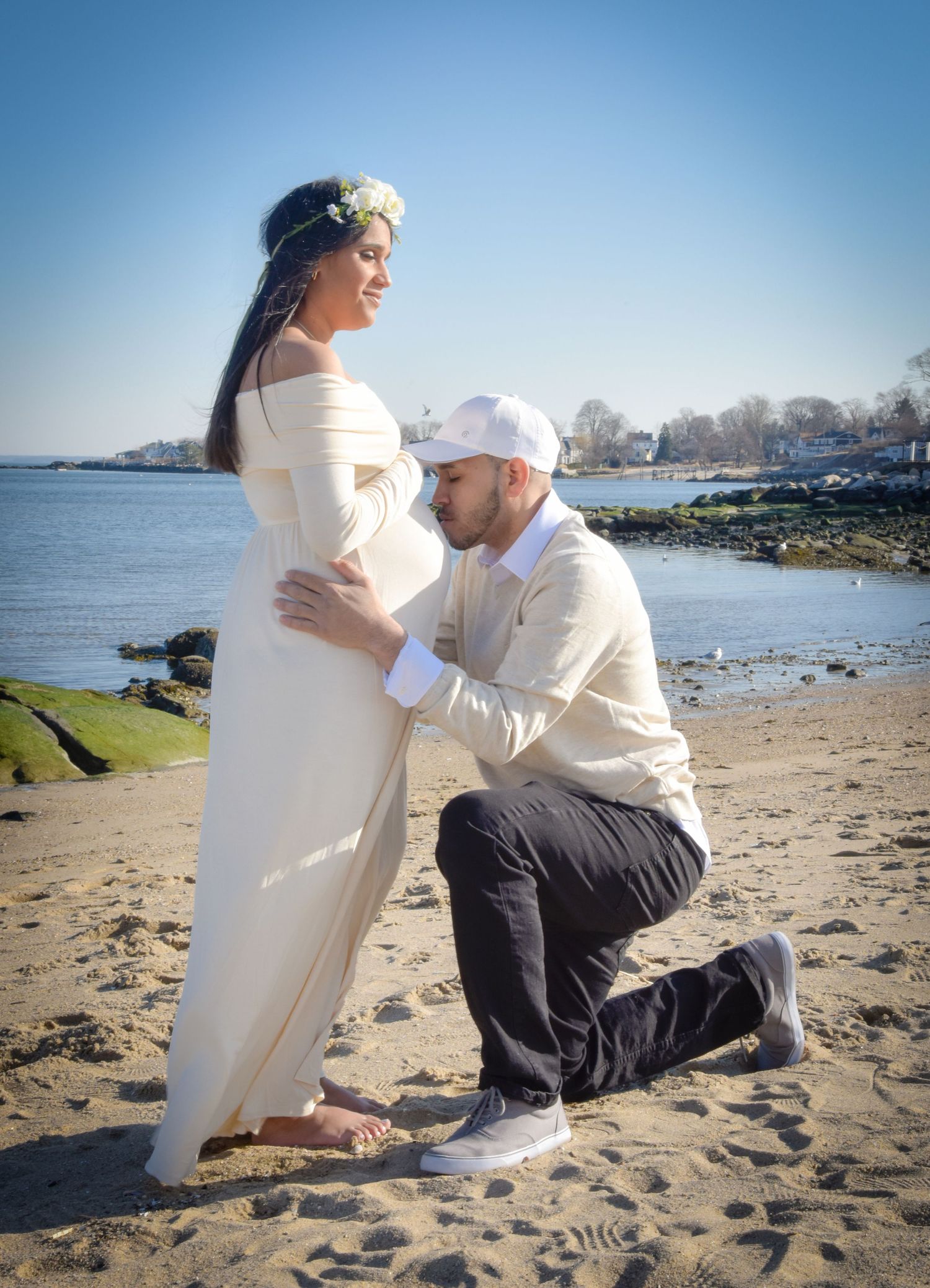 A romantic maternity photo shoot on a beach with an expectant couple posed by the waterfront on a sunny day.