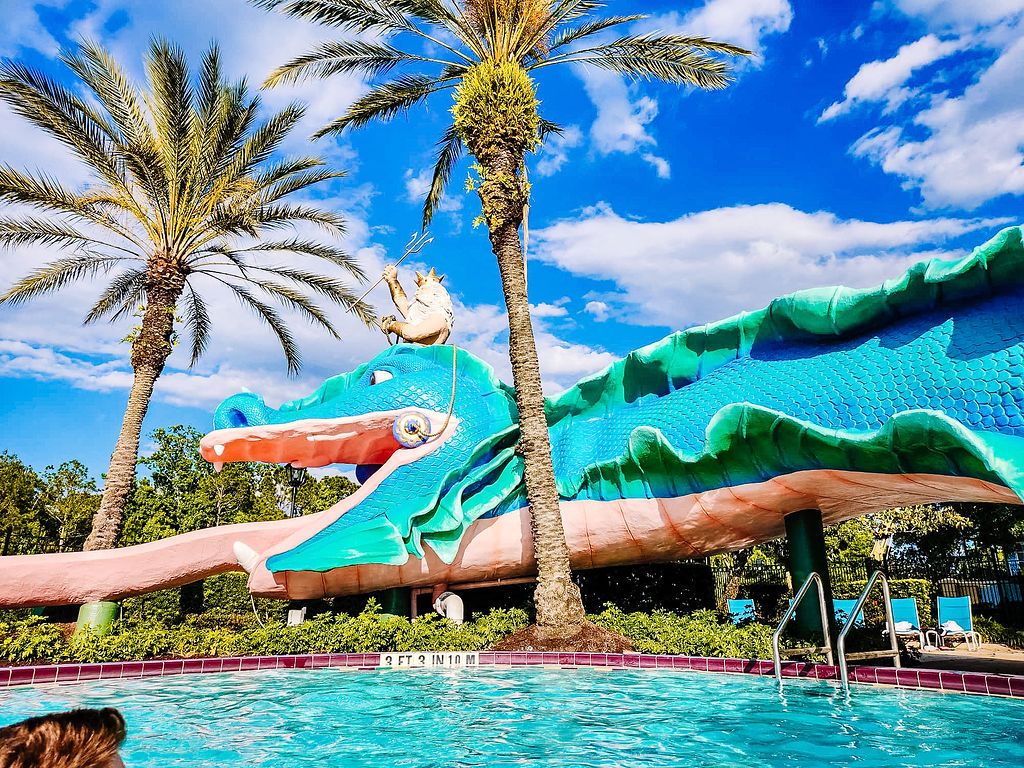 Panoramic view of a resort swimming pool with blue water slides and palm trees against a bright blue sky.