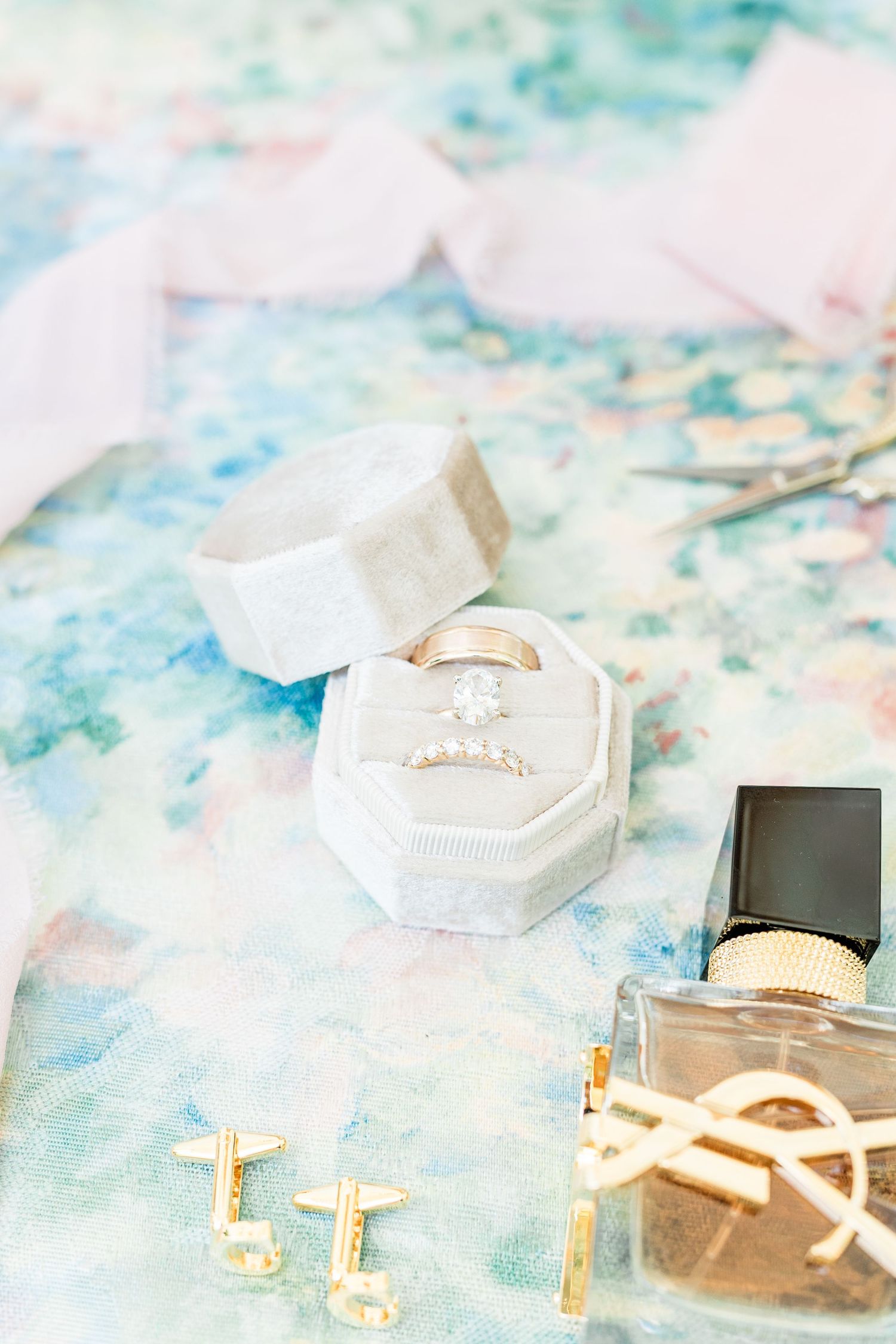 Wedding accessories including engagement ring in velvet box, gold earrings and perfume bottle on pastel watercolor background.