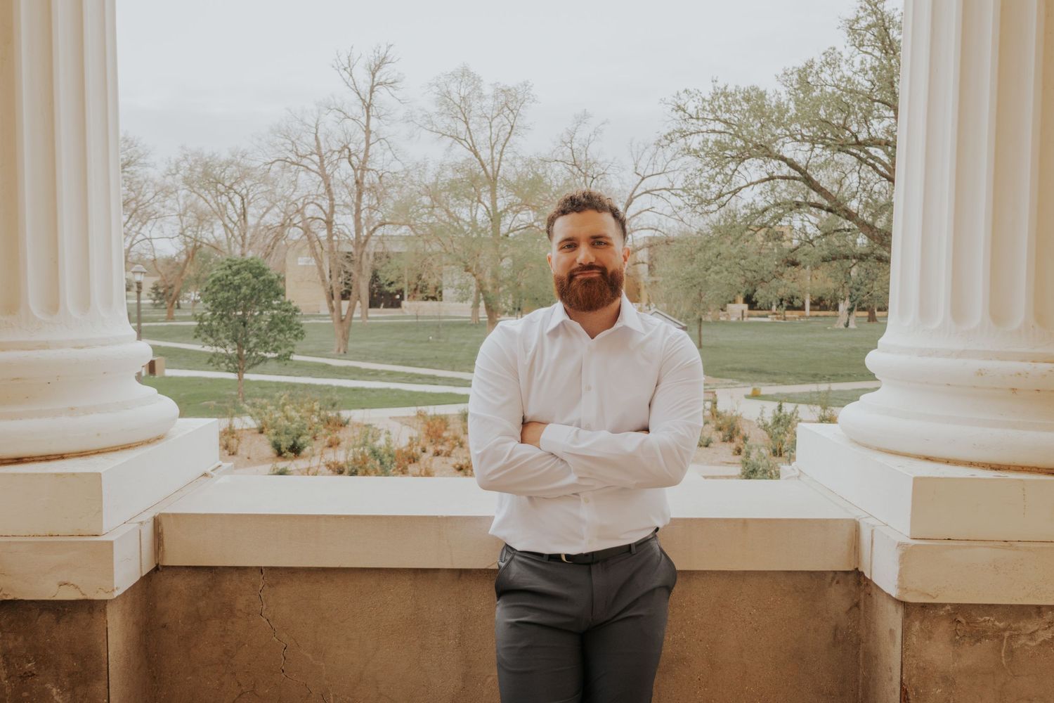 A person stands between white columns on a stone balcony overlooking a park landscape at sunset.