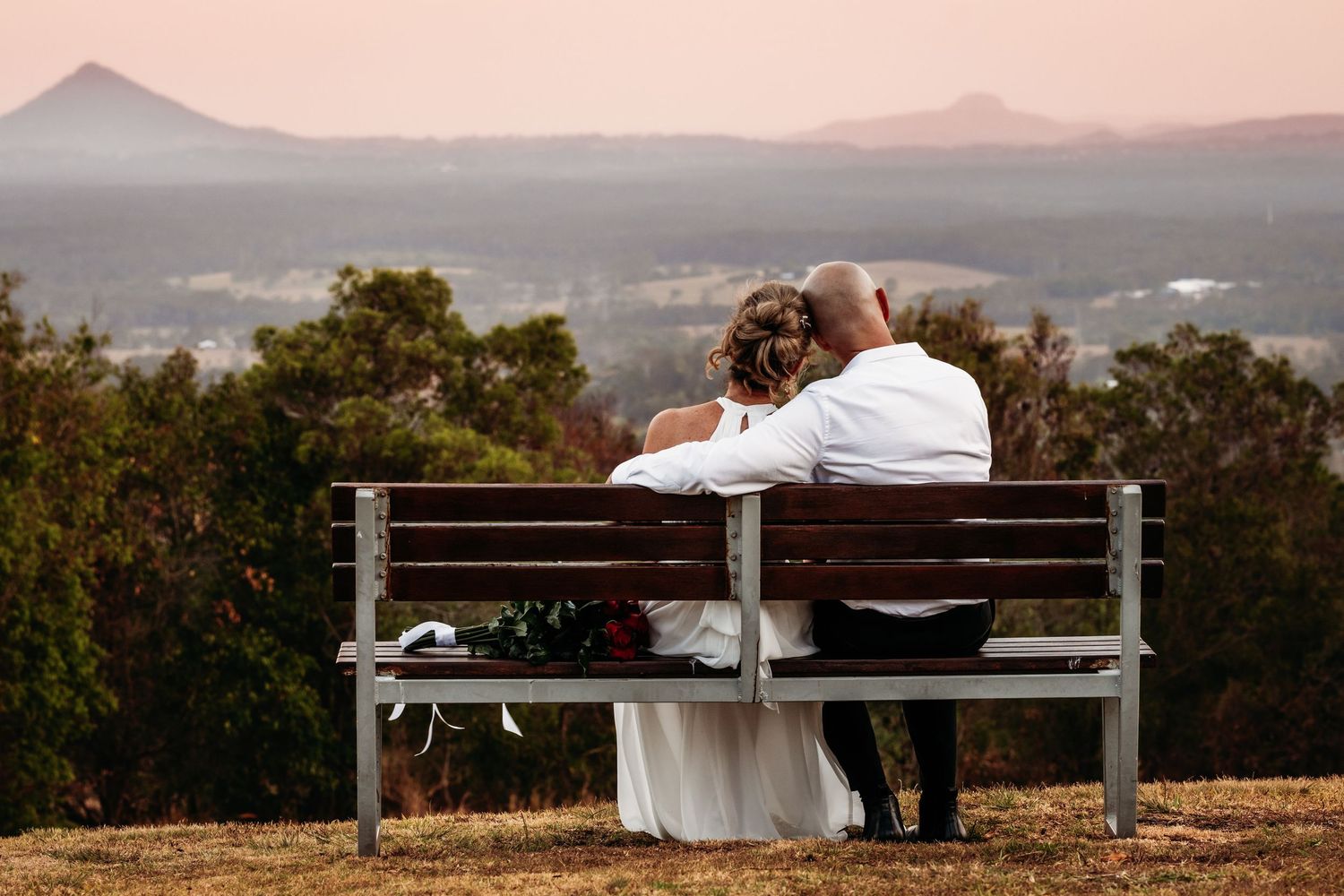 A couple sits together on a wooden bench overlooking mountain vistas at sunset creating a romantic silhouette.