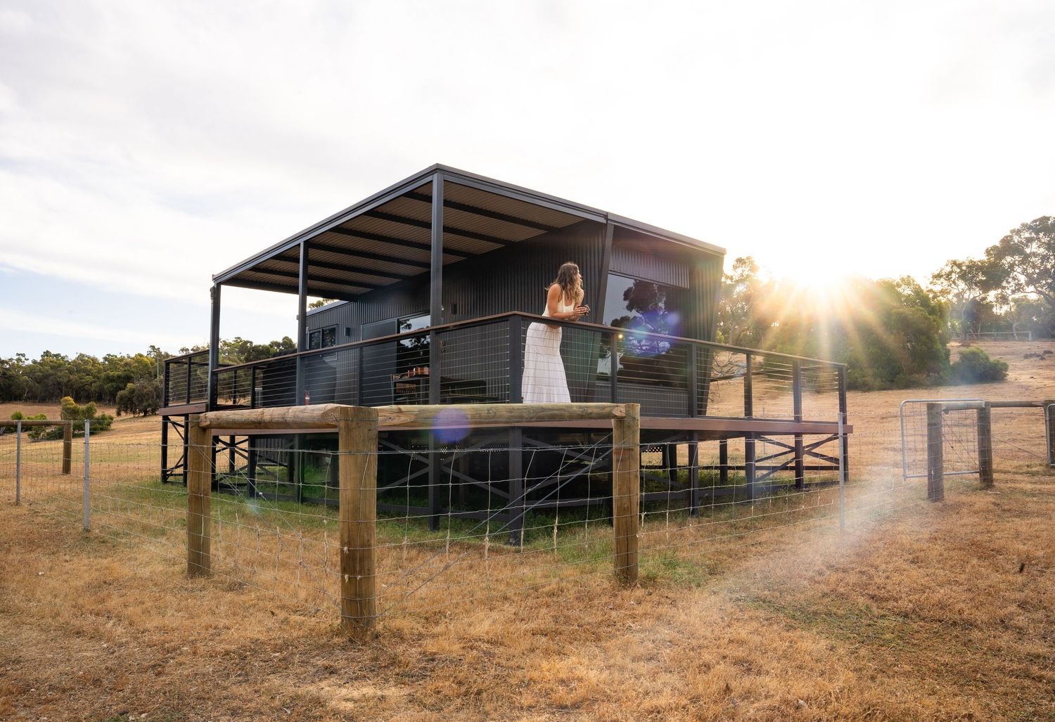 Modern elevated cabin with metal roof and deck overlooking a rural landscape at sunset.