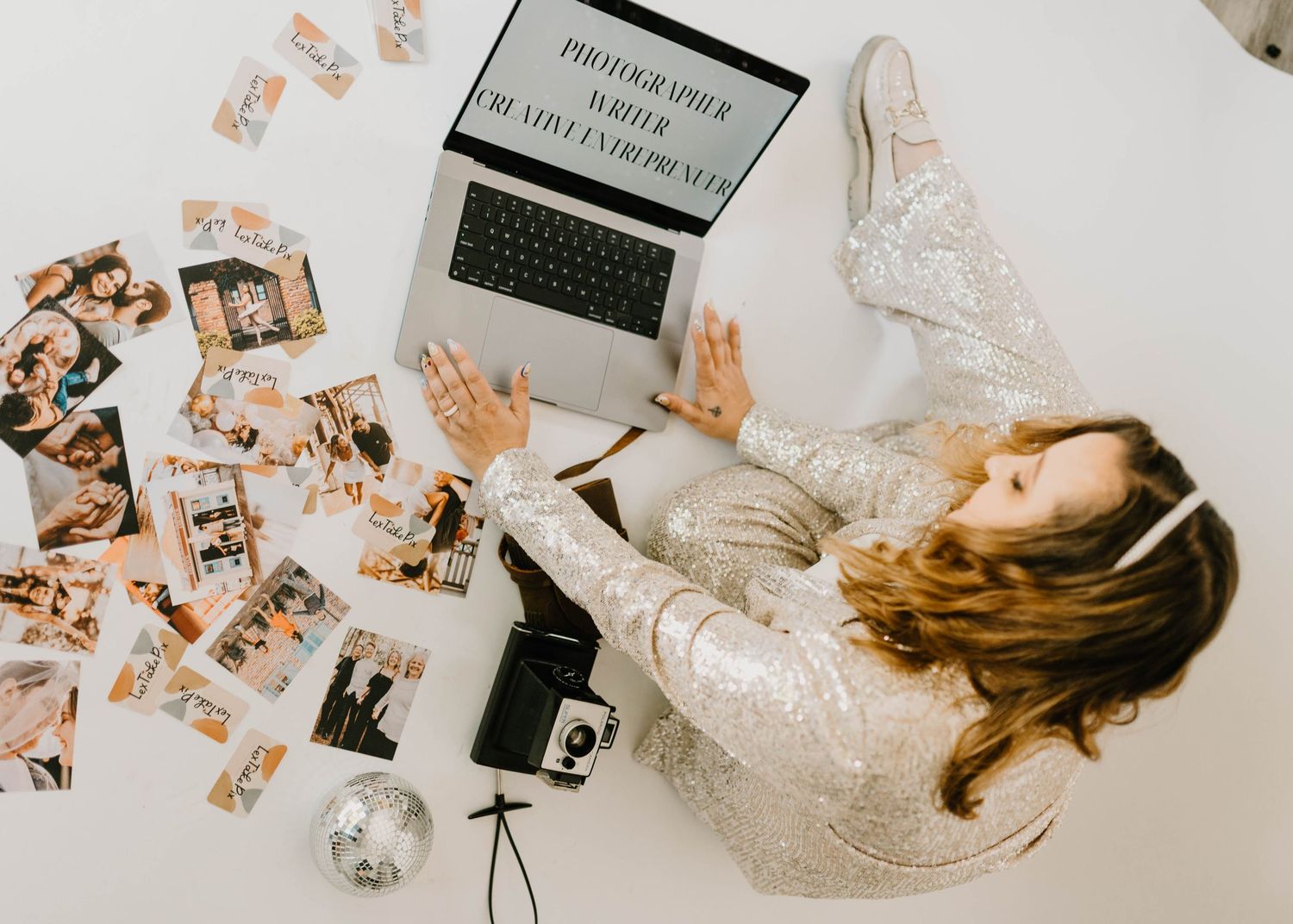 Alexis Stuart sitting on studio floor on her computer with photos & business cards spread out, wearing a Nine West sequin suit. Cred: Angela