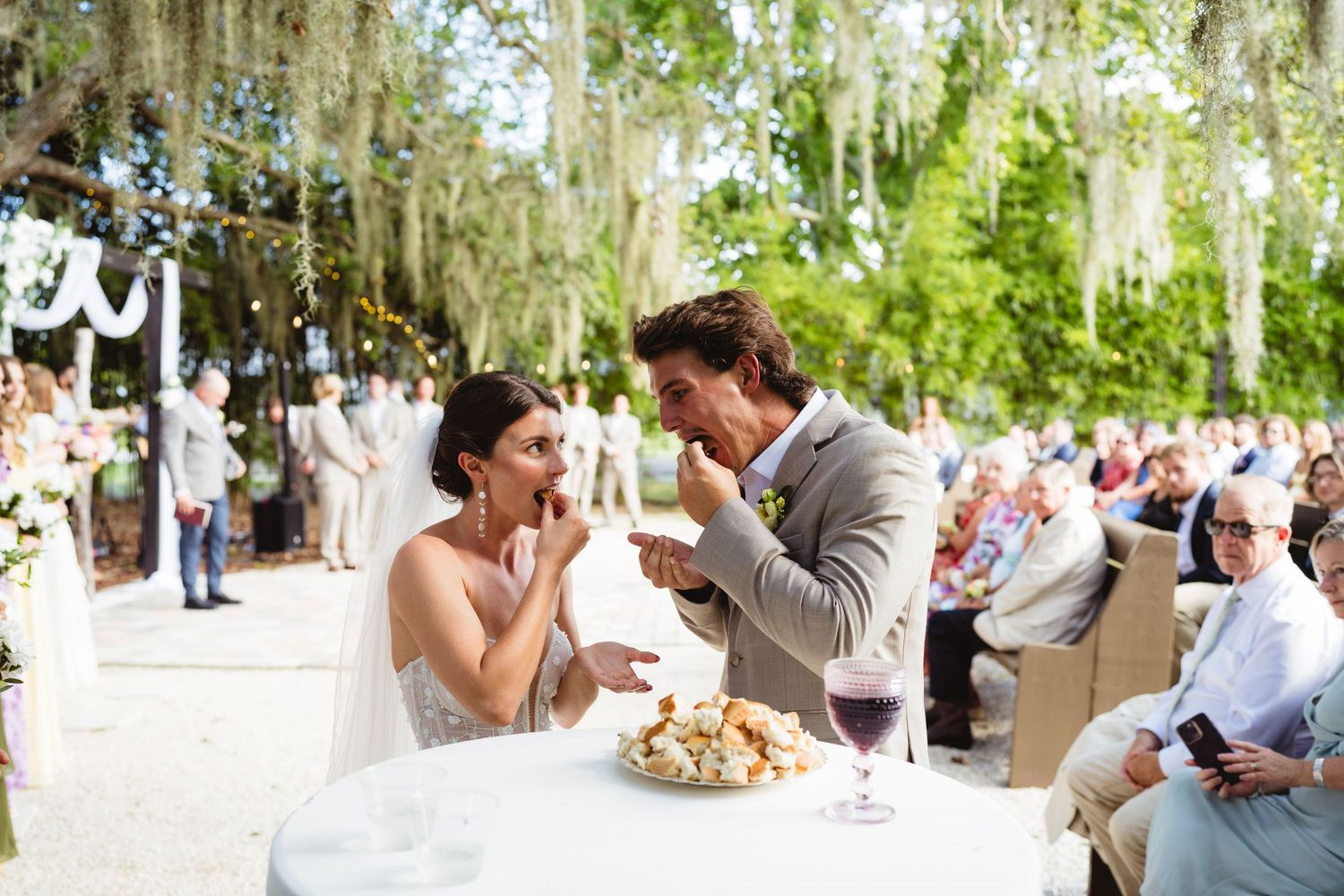 Newlyweds share a joyful moment feeding each other cake at an outdoor garden wedding reception under hanging moss.