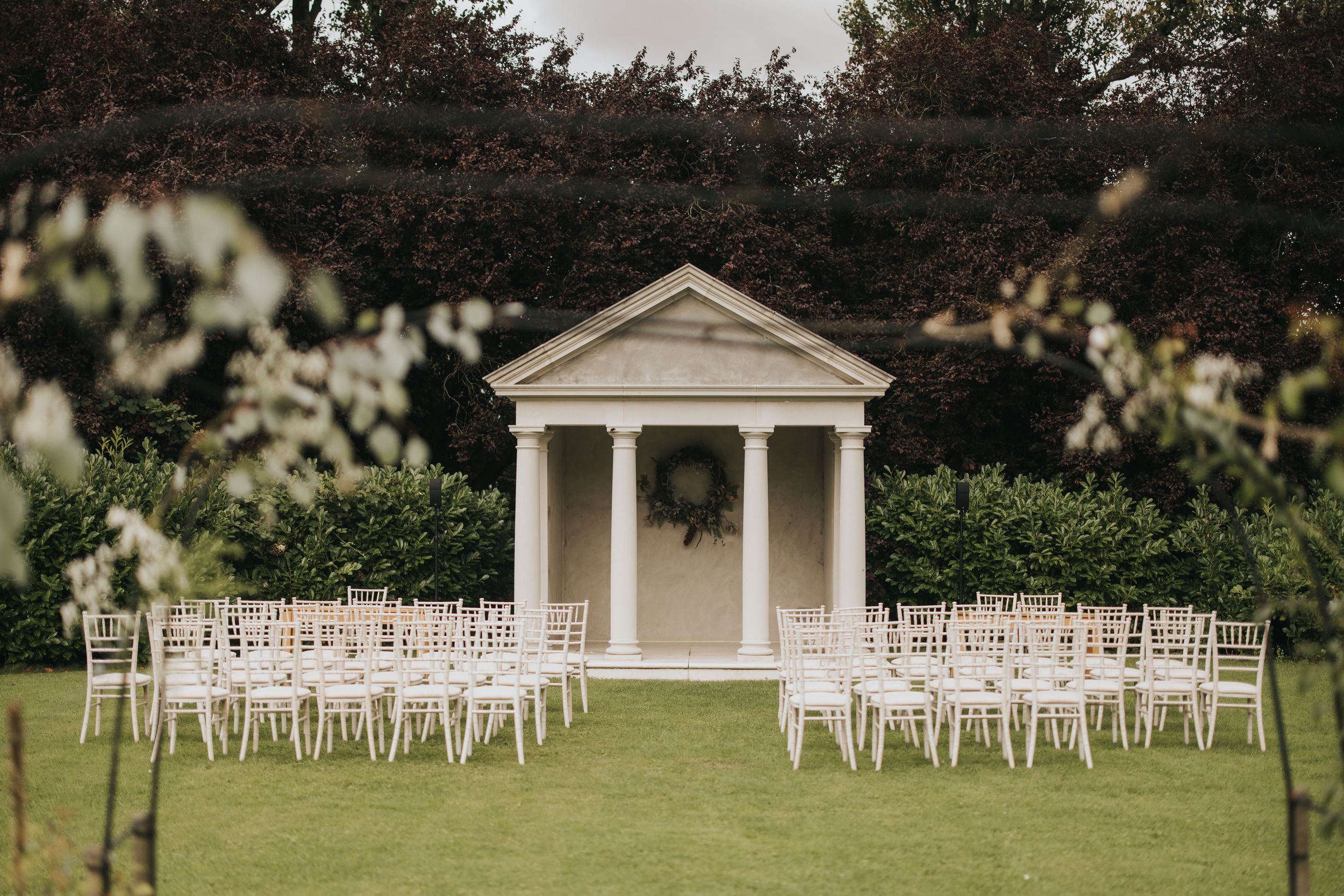White wedding chairs arranged in rows facing a classical columned pavilion on a lush green lawn at an outdoor venue.