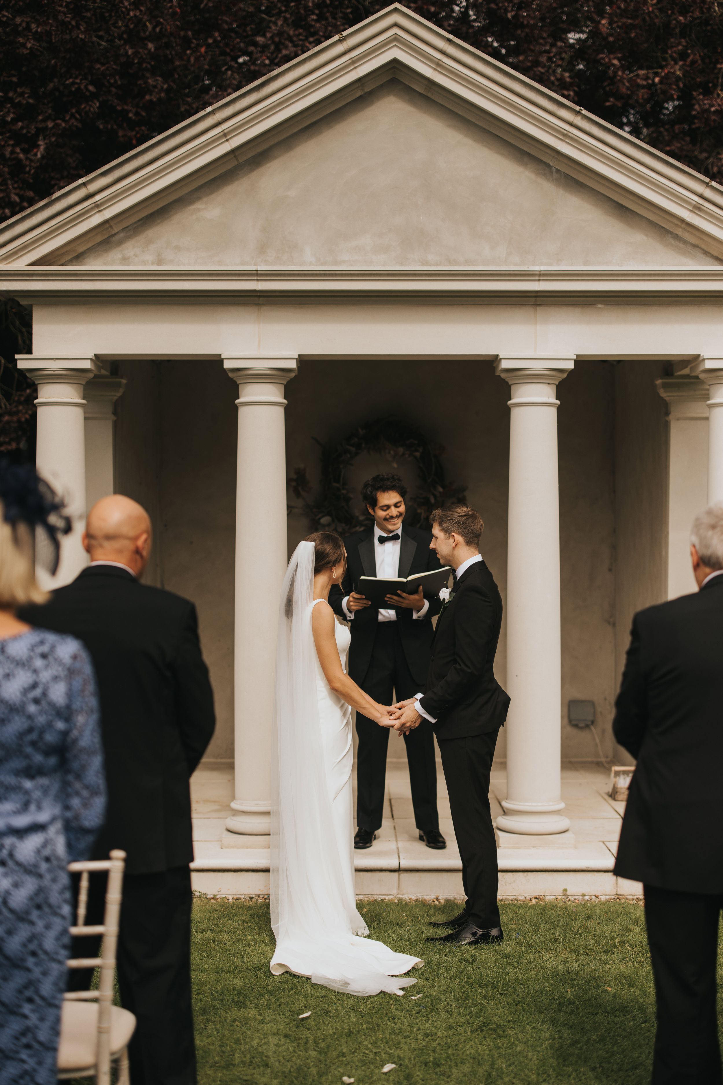 Outdoor wedding ceremony taking place at an elegant white columned gazebo surrounded by trees.