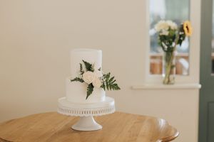 Simple two-tier white wedding cake with floral decorations sits on a white cake stand on wooden table with vase in background.