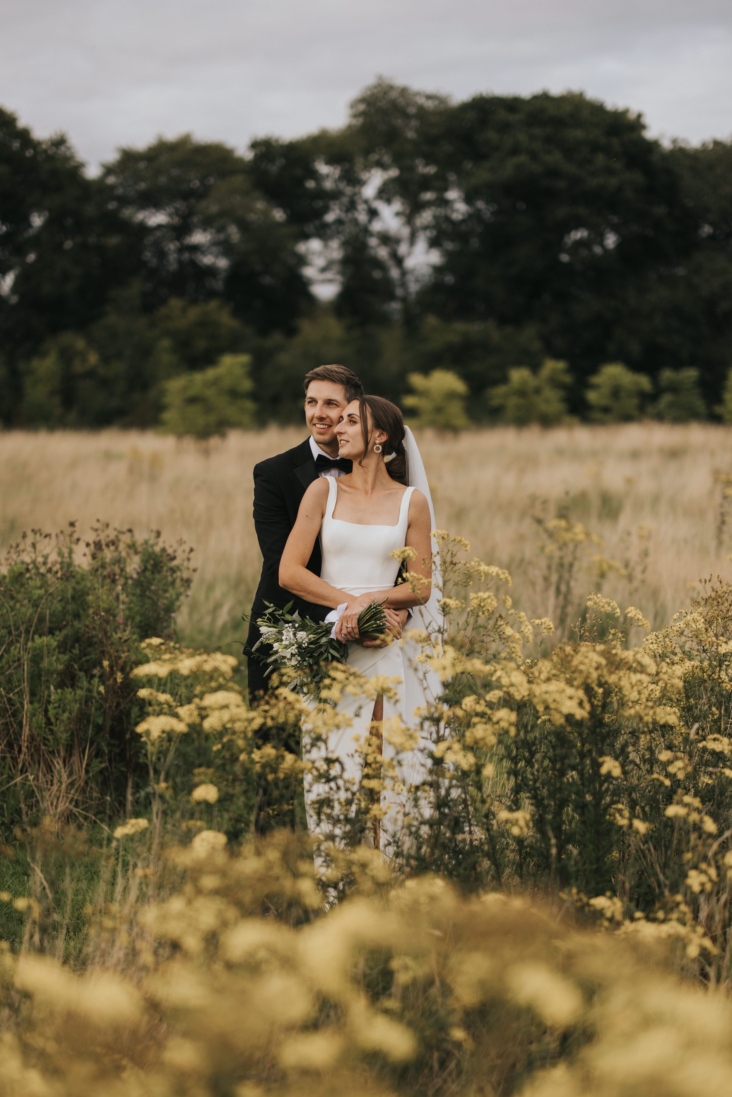 A romantic wedding couple embraces in a sunlit meadow filled with golden wildflowers at sunset.