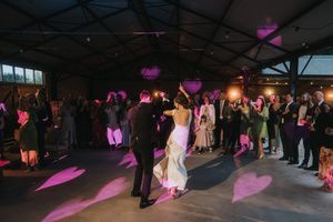 Wedding reception guests dance in a large industrial venue with purple spotlight accents on the dance floor.