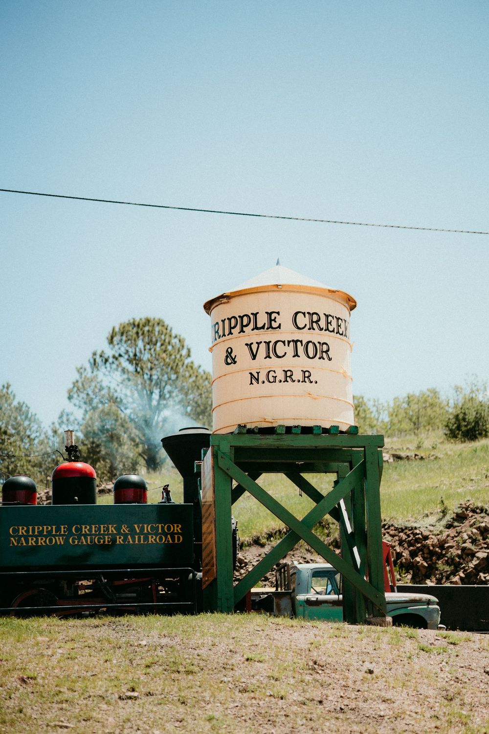 A vintage train water tower marked 'Cripple Creek & Victor N.G.R.R.' stands on wooden supports against a blue sky.