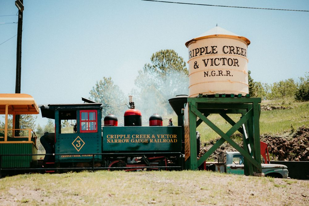 A vintage green steam train passes under a wooden water tower marked 'Apple Creek & Victor' at a historic railway station.
