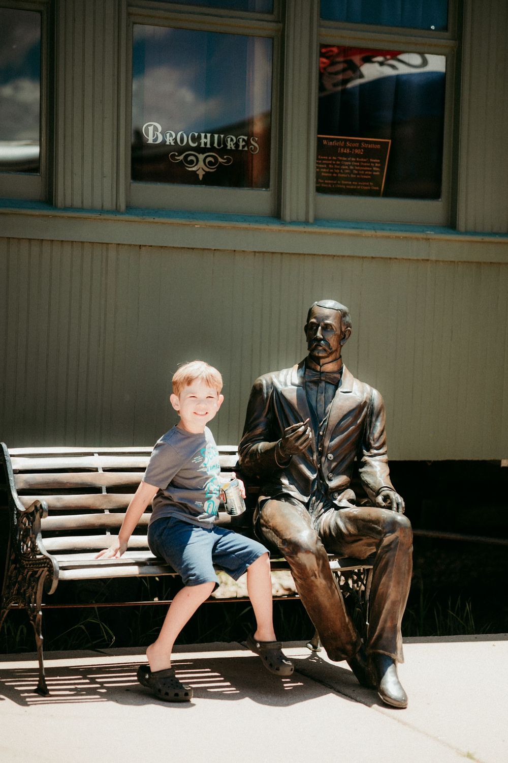 A child sits next to a bronze statue on a wooden bench outside a storefront with a 'Brochures' sign in the window.