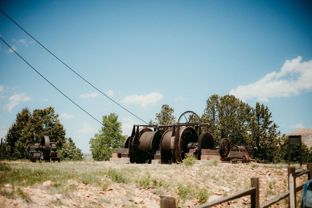 Vintage rustic farm equipment sits abandoned in a rural field against a bright blue summer sky with trees in background.
