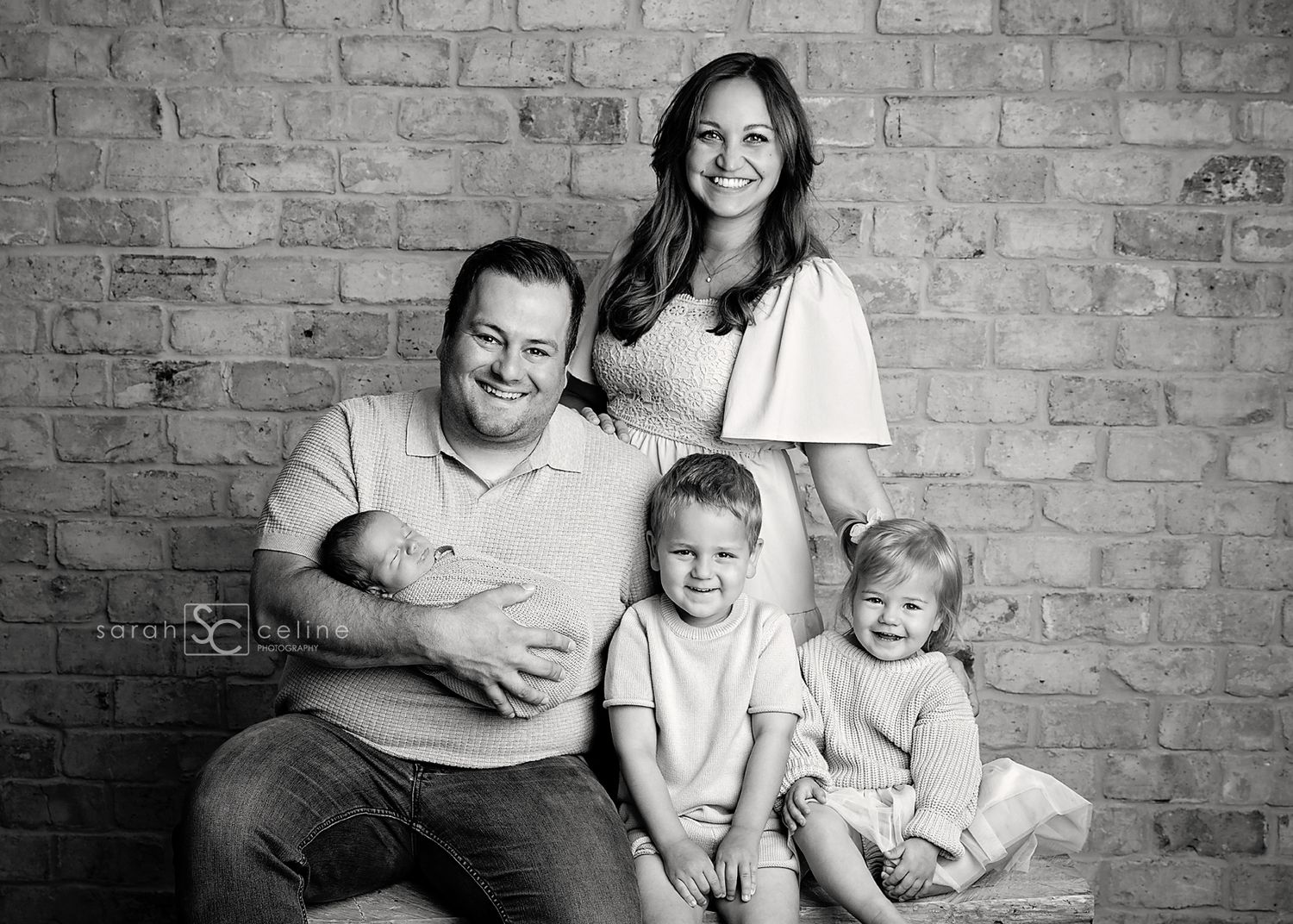 Black and white portrait of a loving family of four posing together against a rustic brick wall.